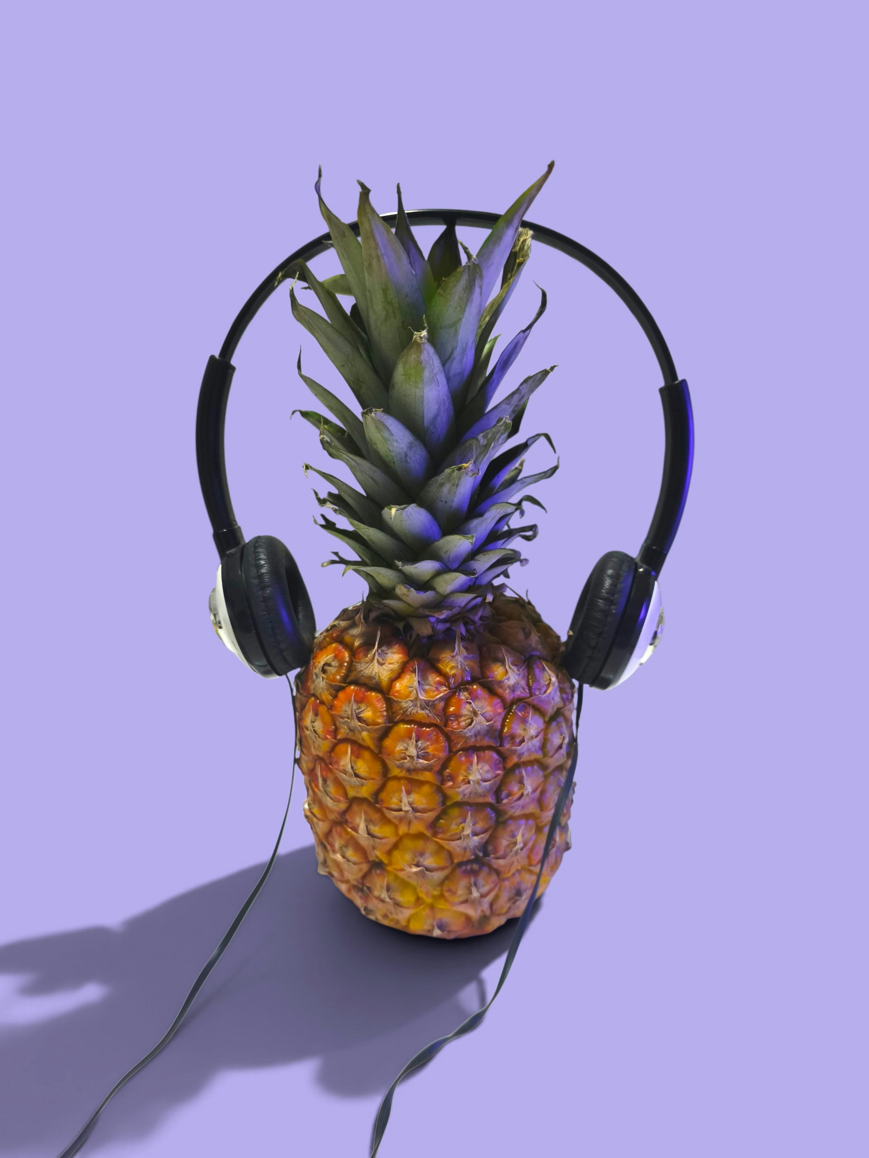 A whole pineapple wearing black headphones against a solid light purple background