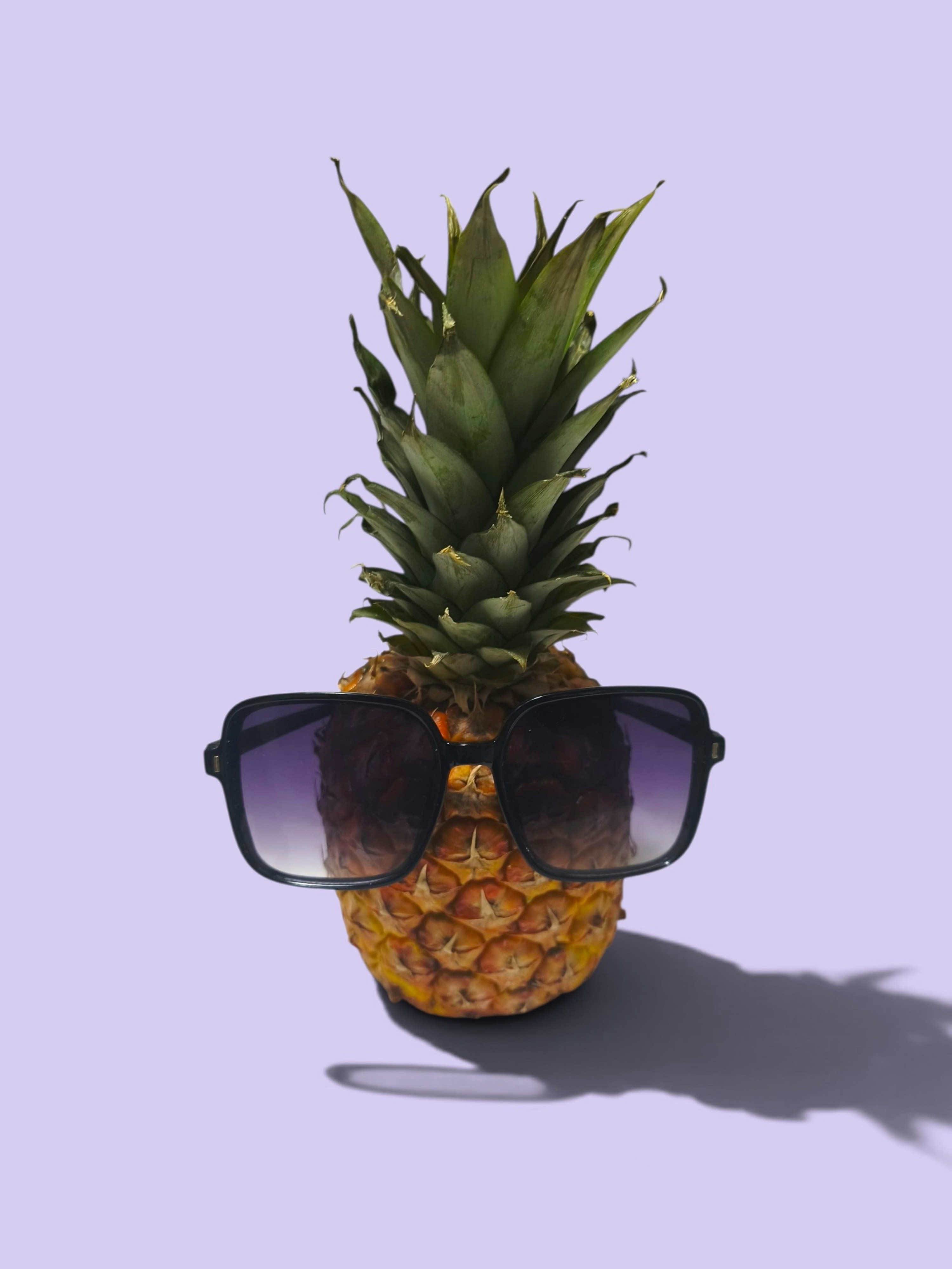 A pineapple wearing large black sunglasses against a solid light purple background