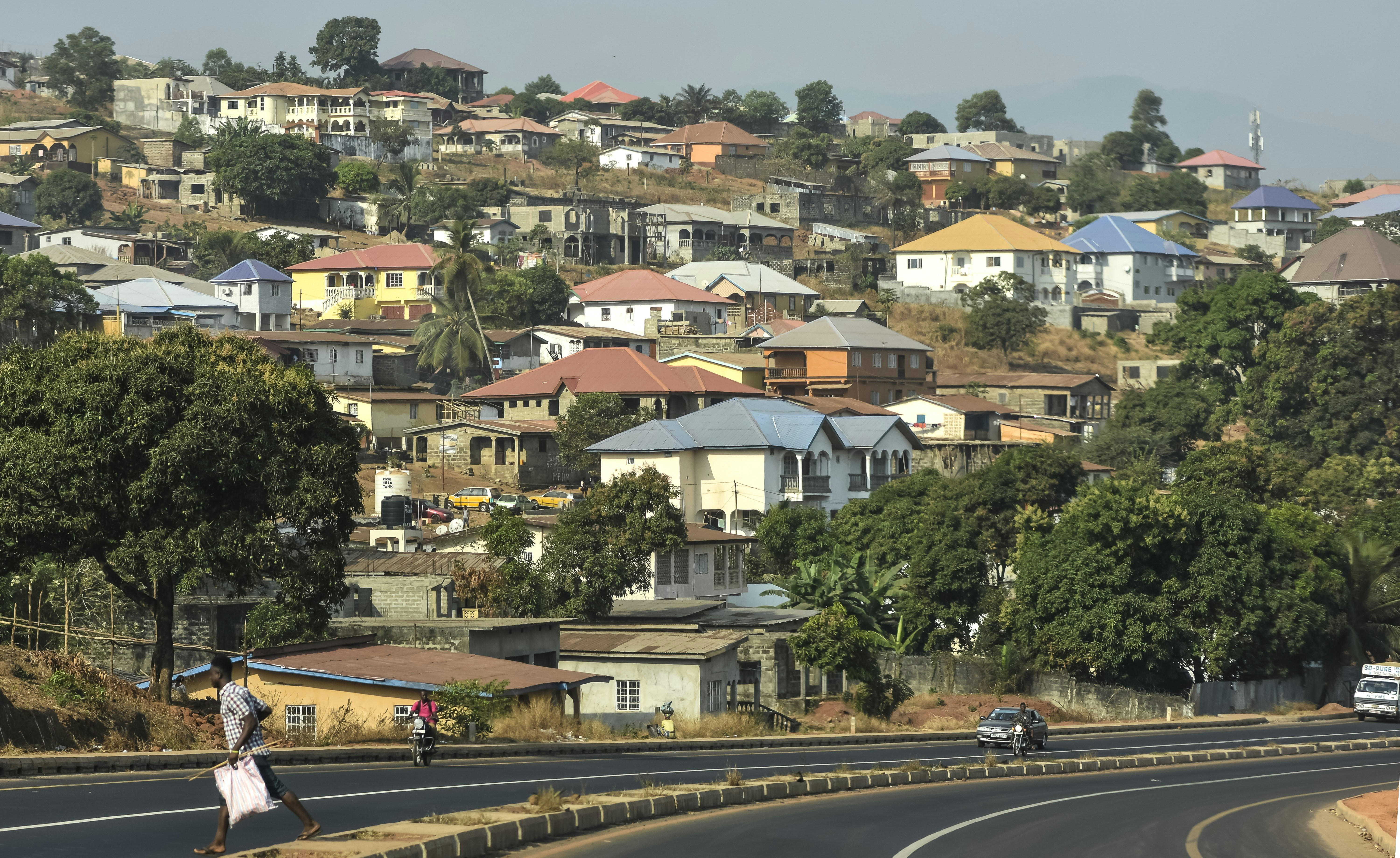 Uma pessoa caminhando por uma rodovia abaixo de uma encosta coberta por casas residenciais