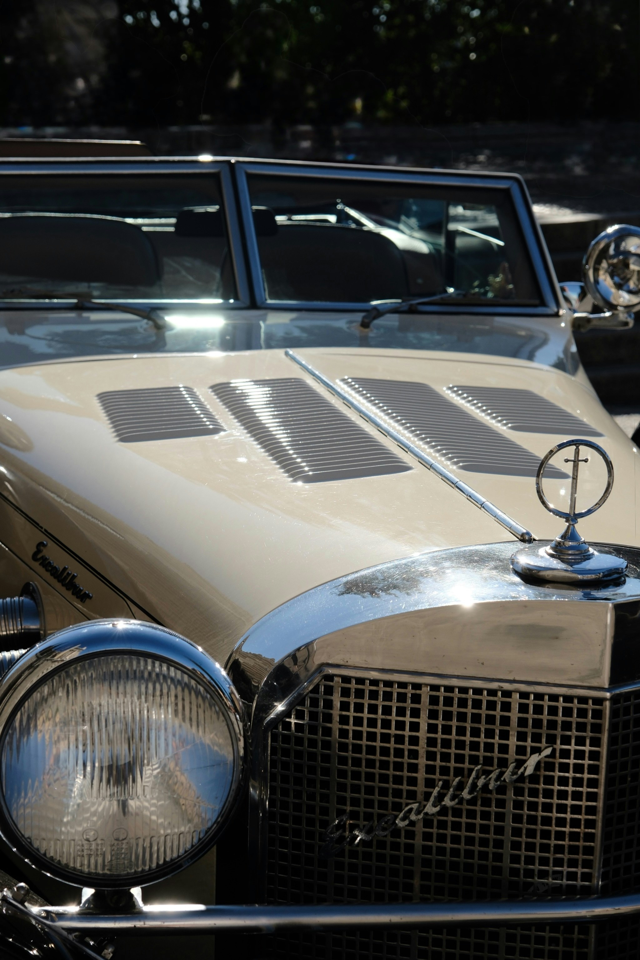 A cream colored vintage Excalibur car with a chrome grille and round headlight