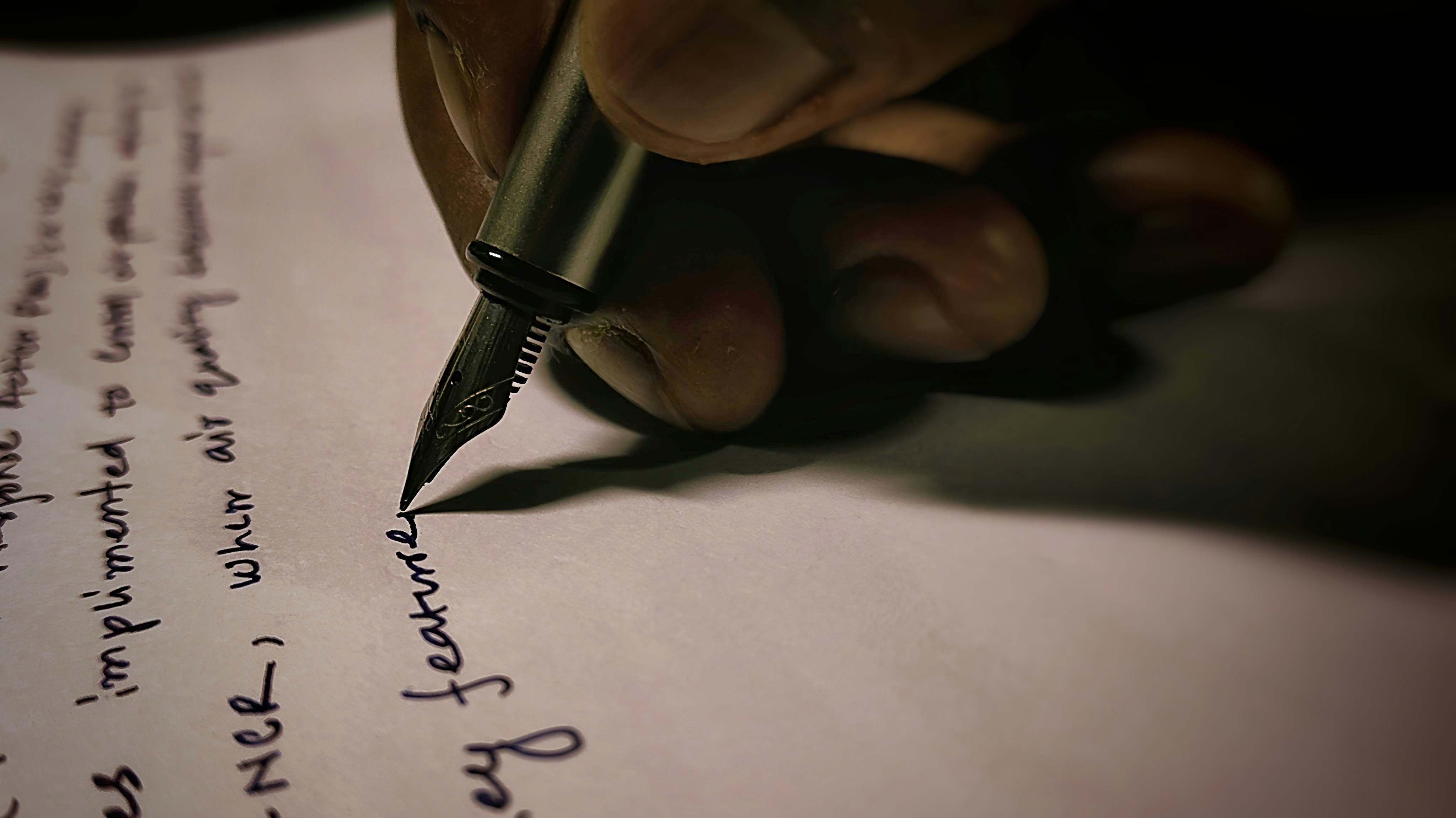 A hand holding a fountain pen writing on white paper in dim light