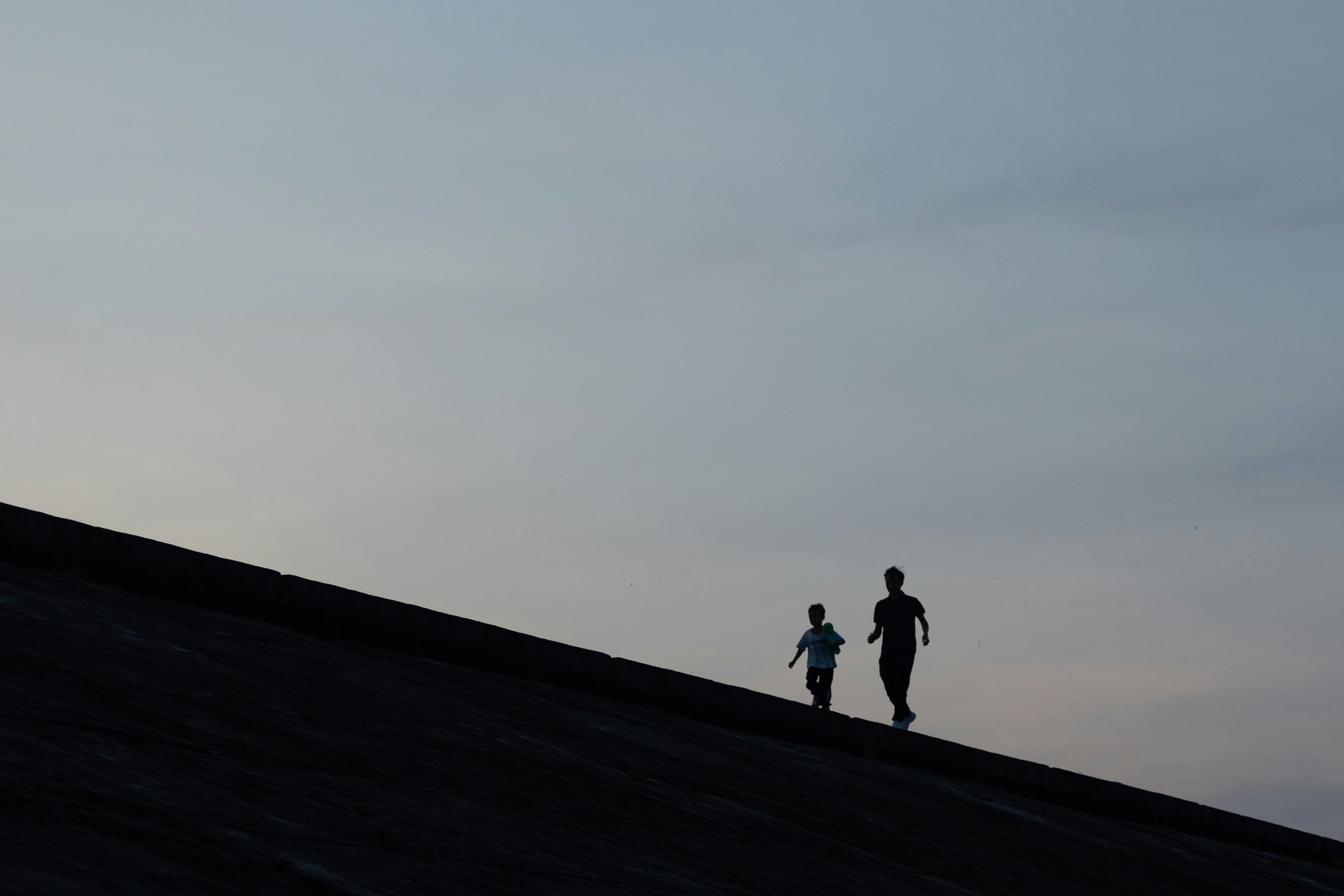 Two people in silhouette walking along a sloped concrete wall against a pale sky