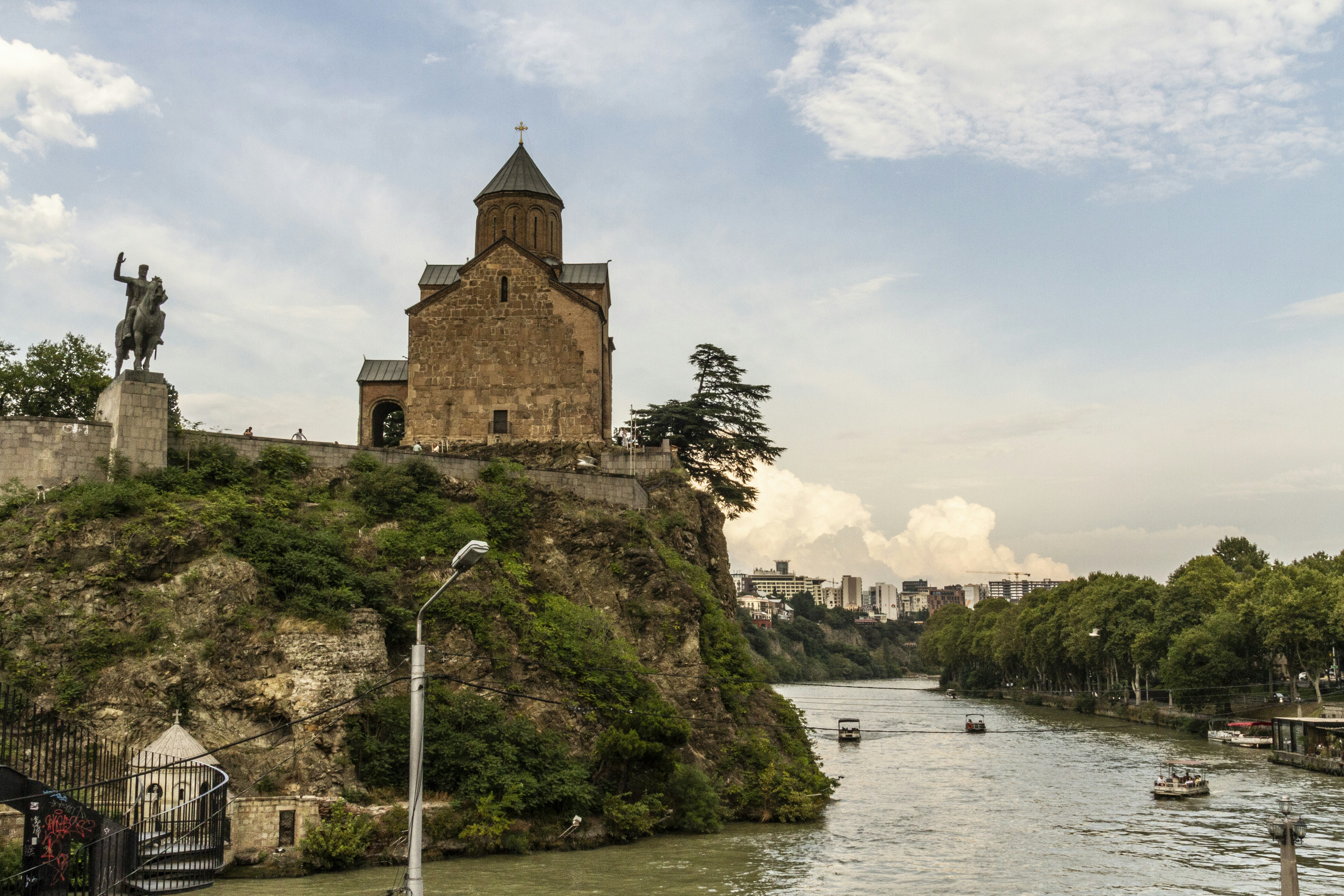A stone church on a cliff above a river with boats in Tbilisi