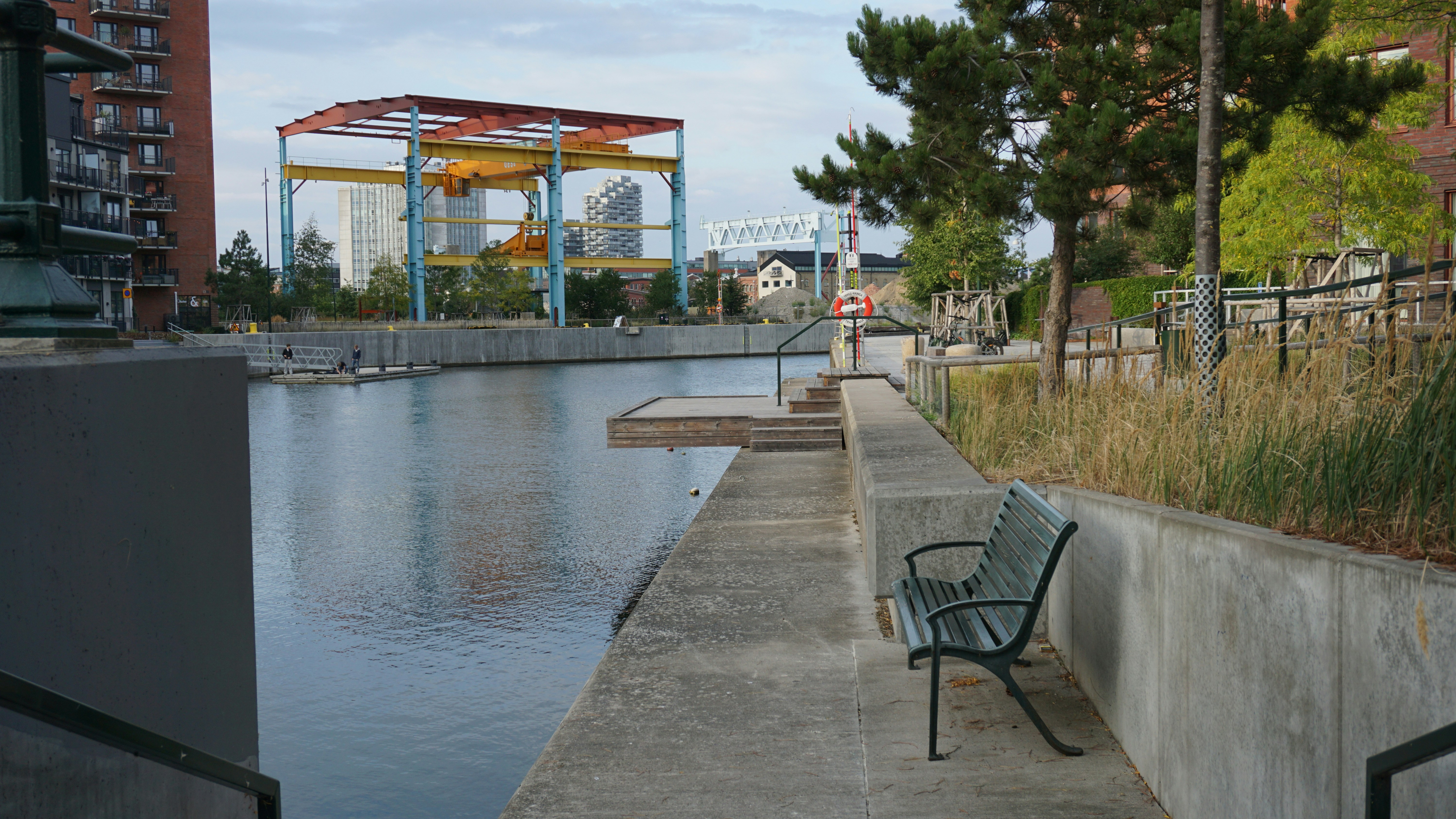 A green bench on a concrete waterfront path beside a canal with industrial cranes