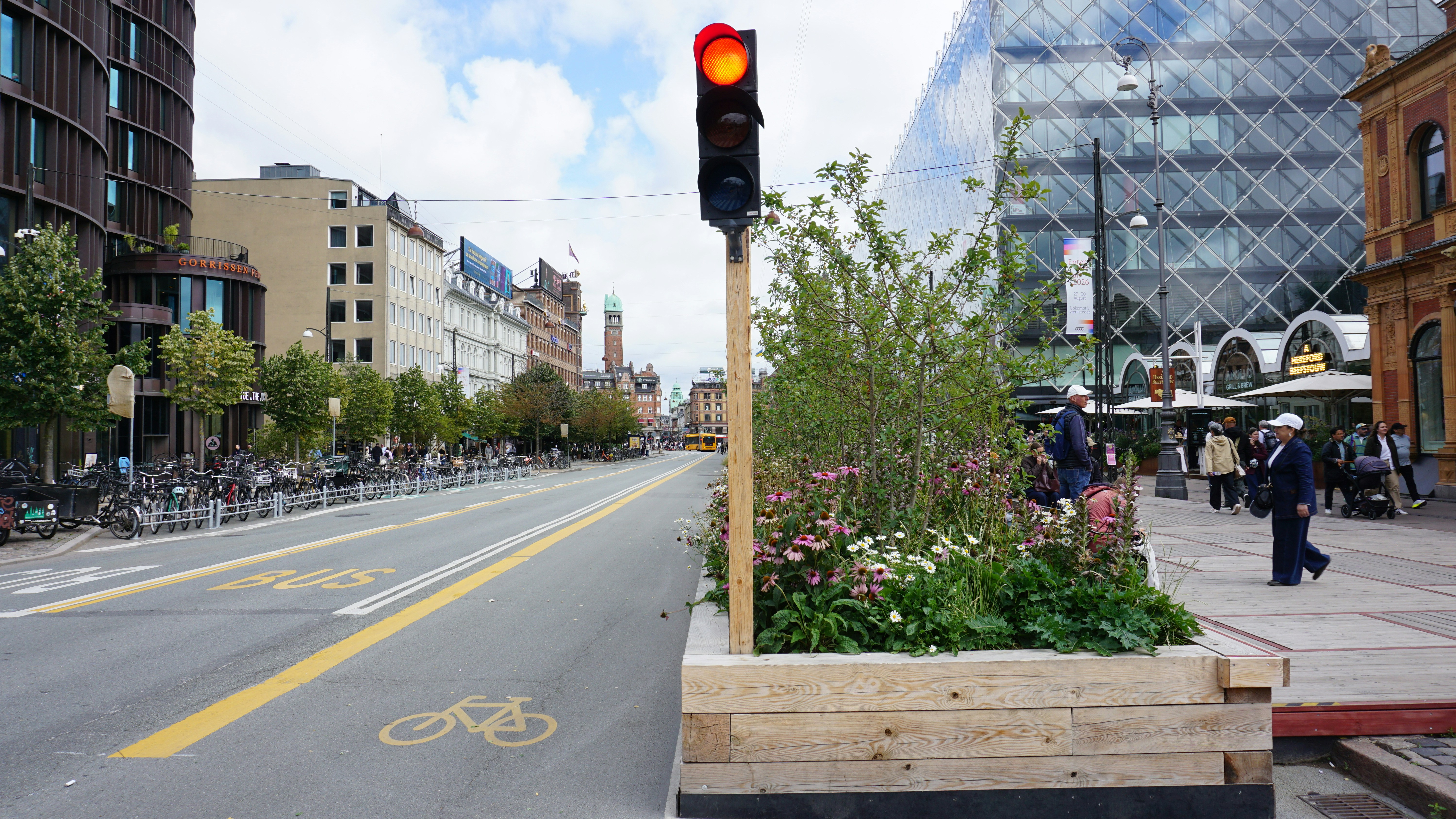 A red traffic light above a wooden planter with flowers on a city street