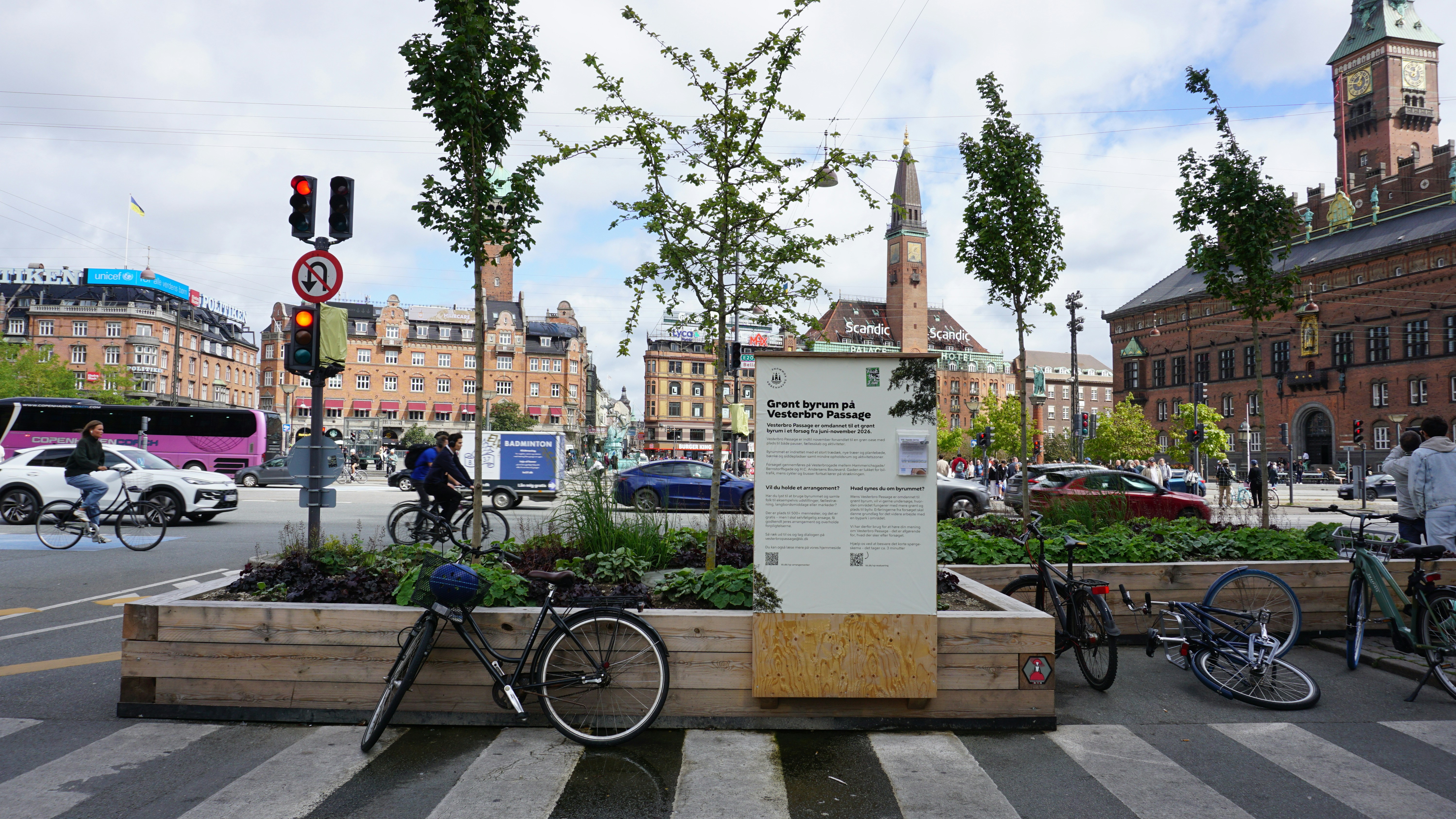 Bicycles parked by a wooden planter with trees in a busy Copenhagen square