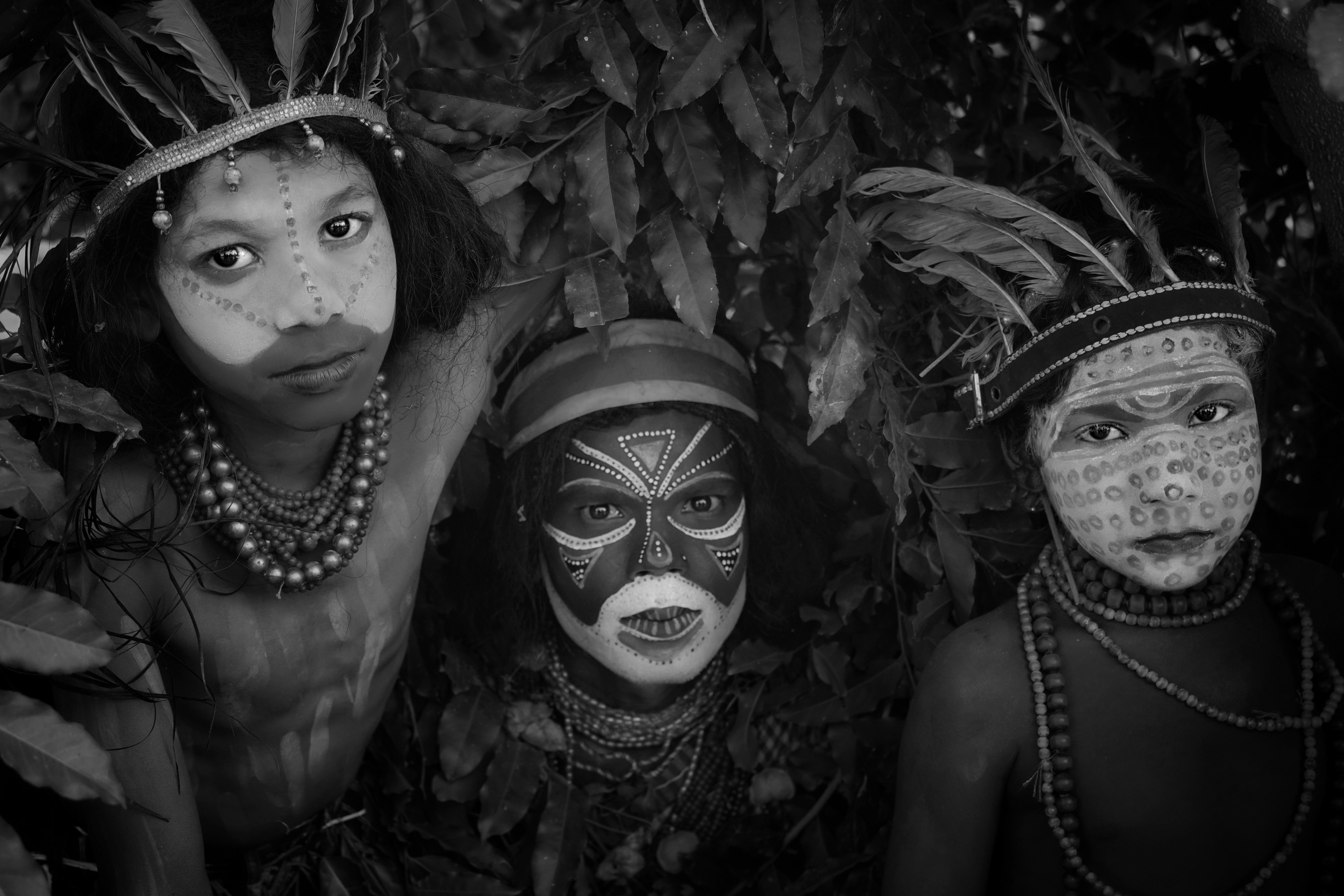 Three people with traditional face paint and feathered headdresses among dense green foliage