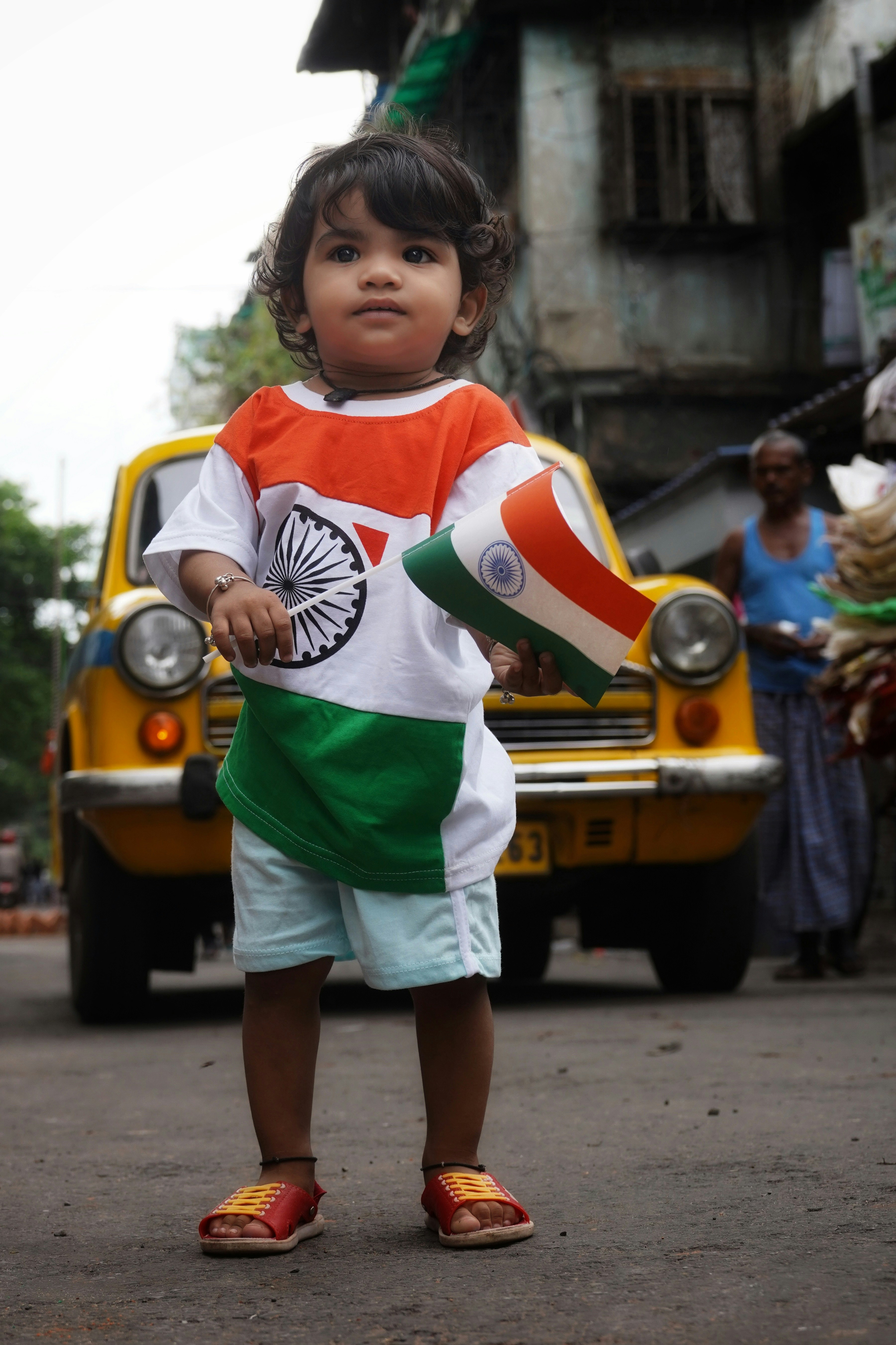 A toddler wearing an Indian flag shirt holding a small flag before a yellow taxi