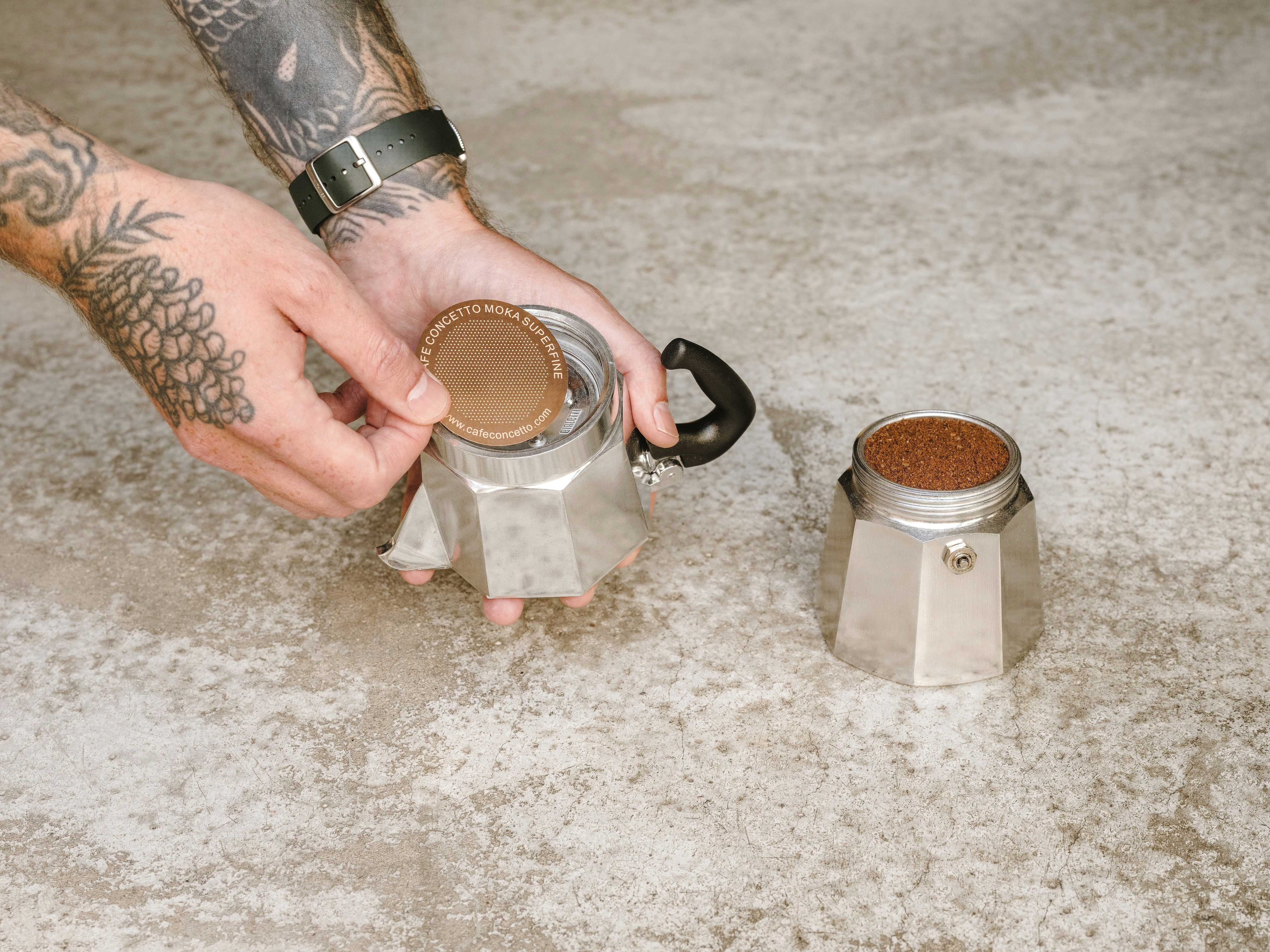 A tattooed person placing a metal filter onto a moka pot filled with coffee grounds