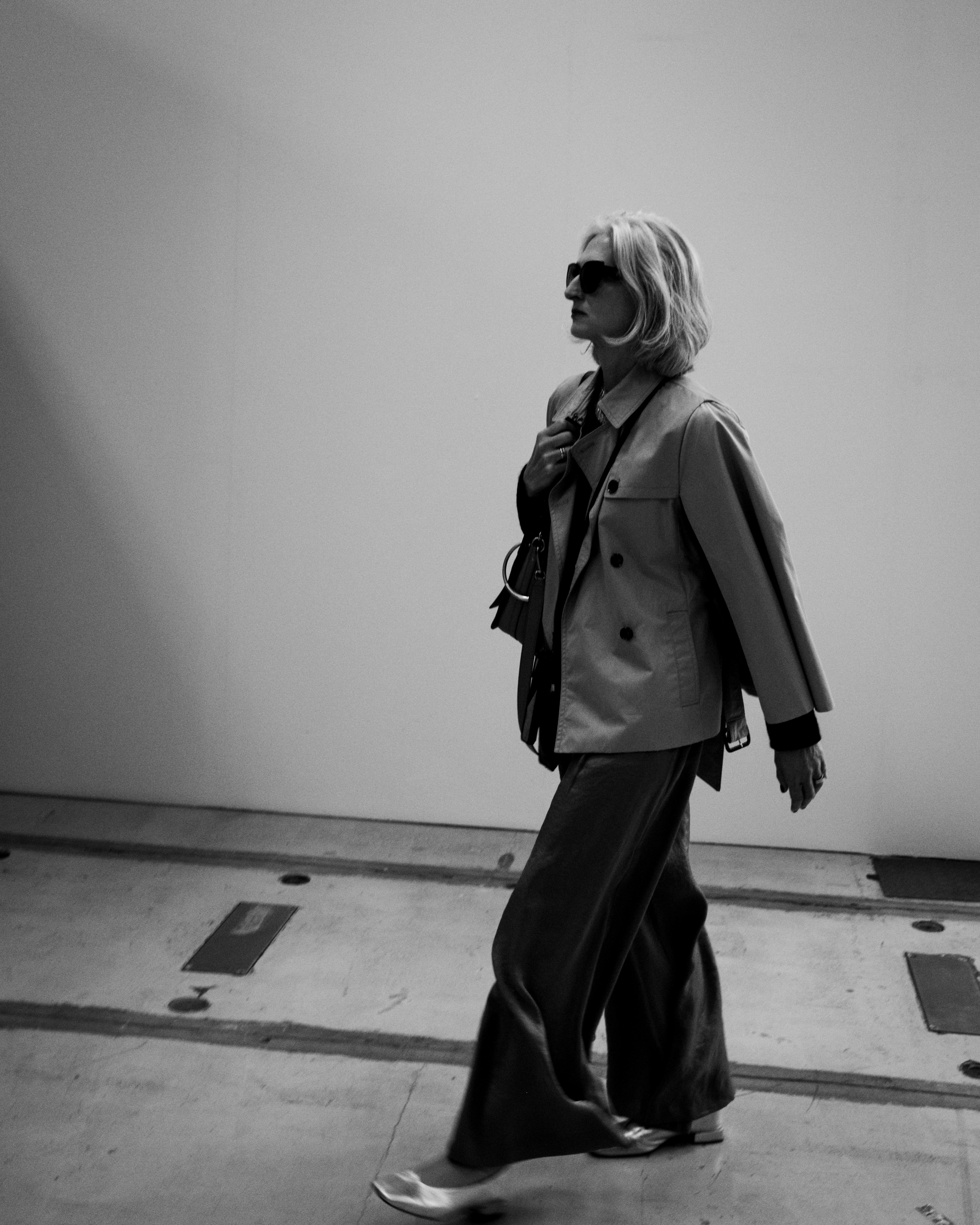 A woman in a trench coat and sunglasses walking across a concrete floor