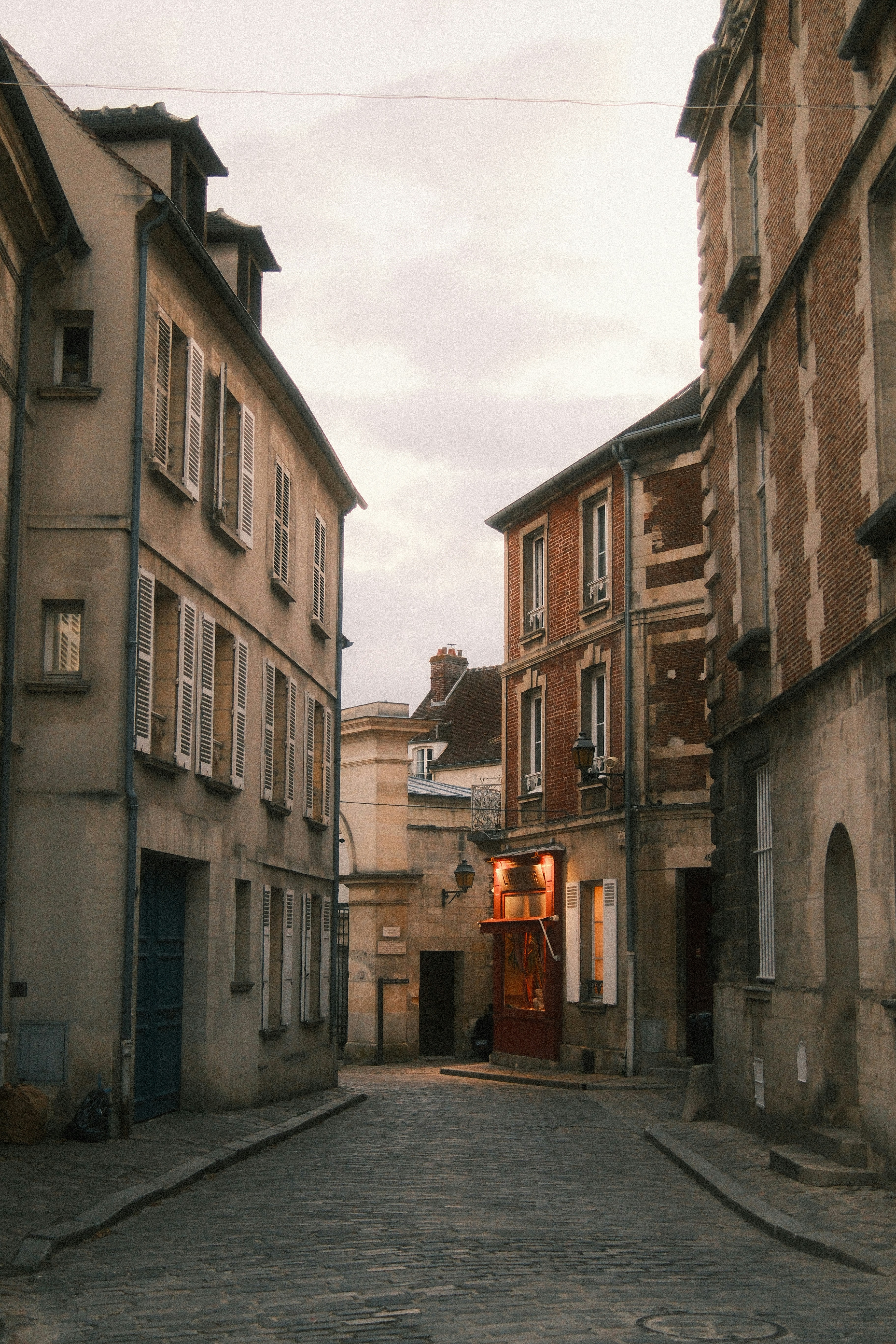 A narrow cobblestone street between historic stone buildings with a small glowing shopfront