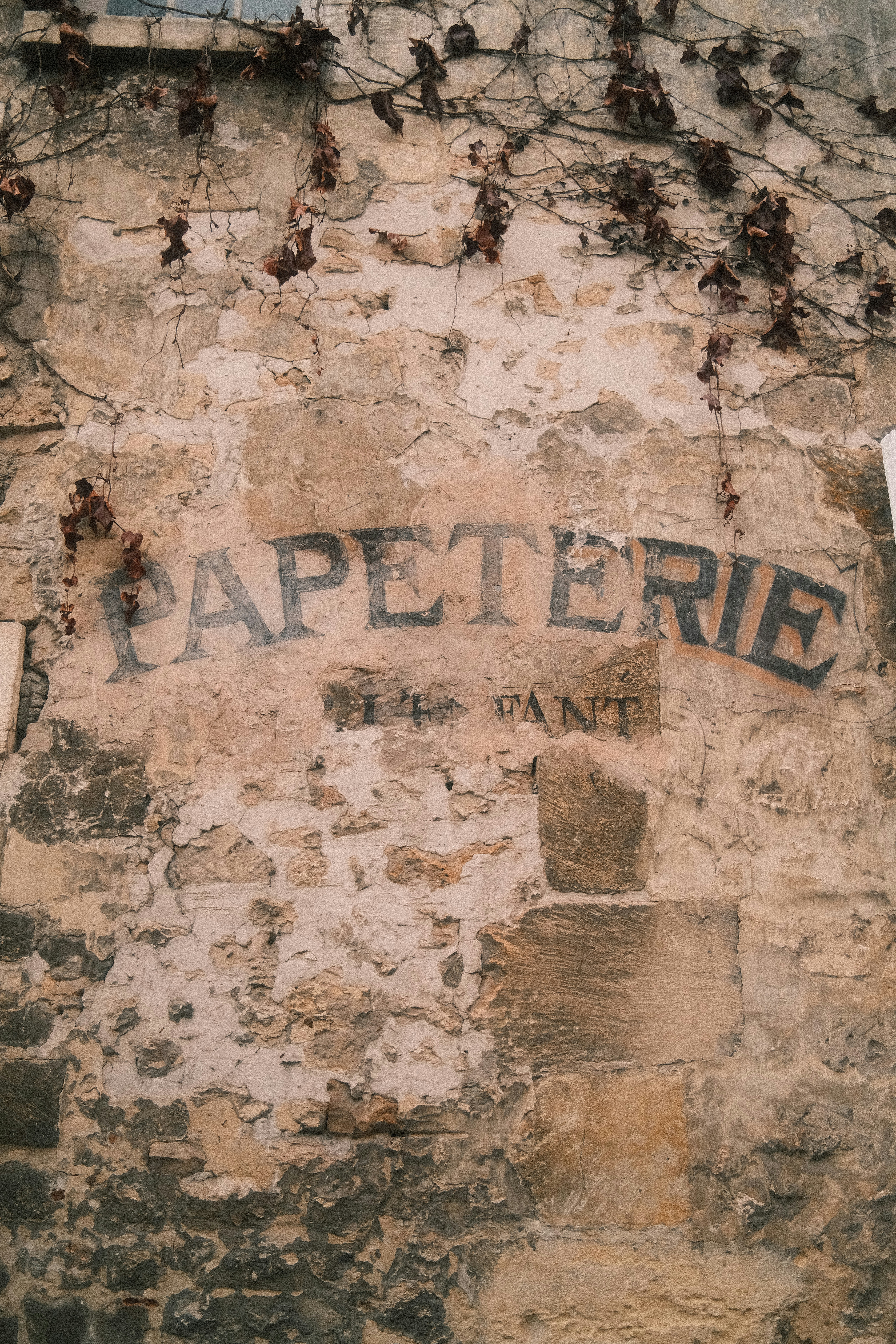 Faded black lettering reading PAPETERIE on a weathered stone wall with dried vines