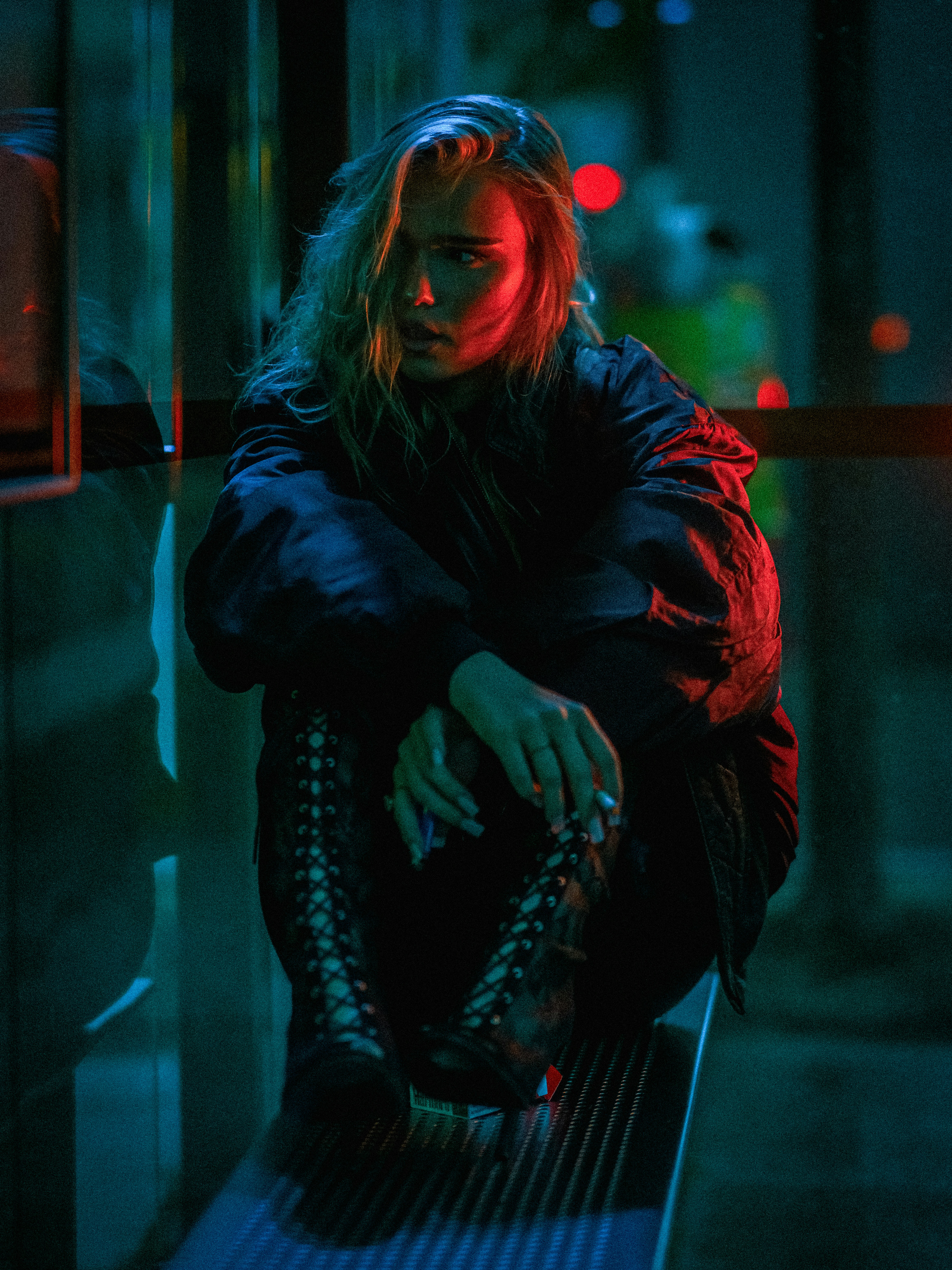 A woman in a dark jacket and lace-up boots sitting under neon lights