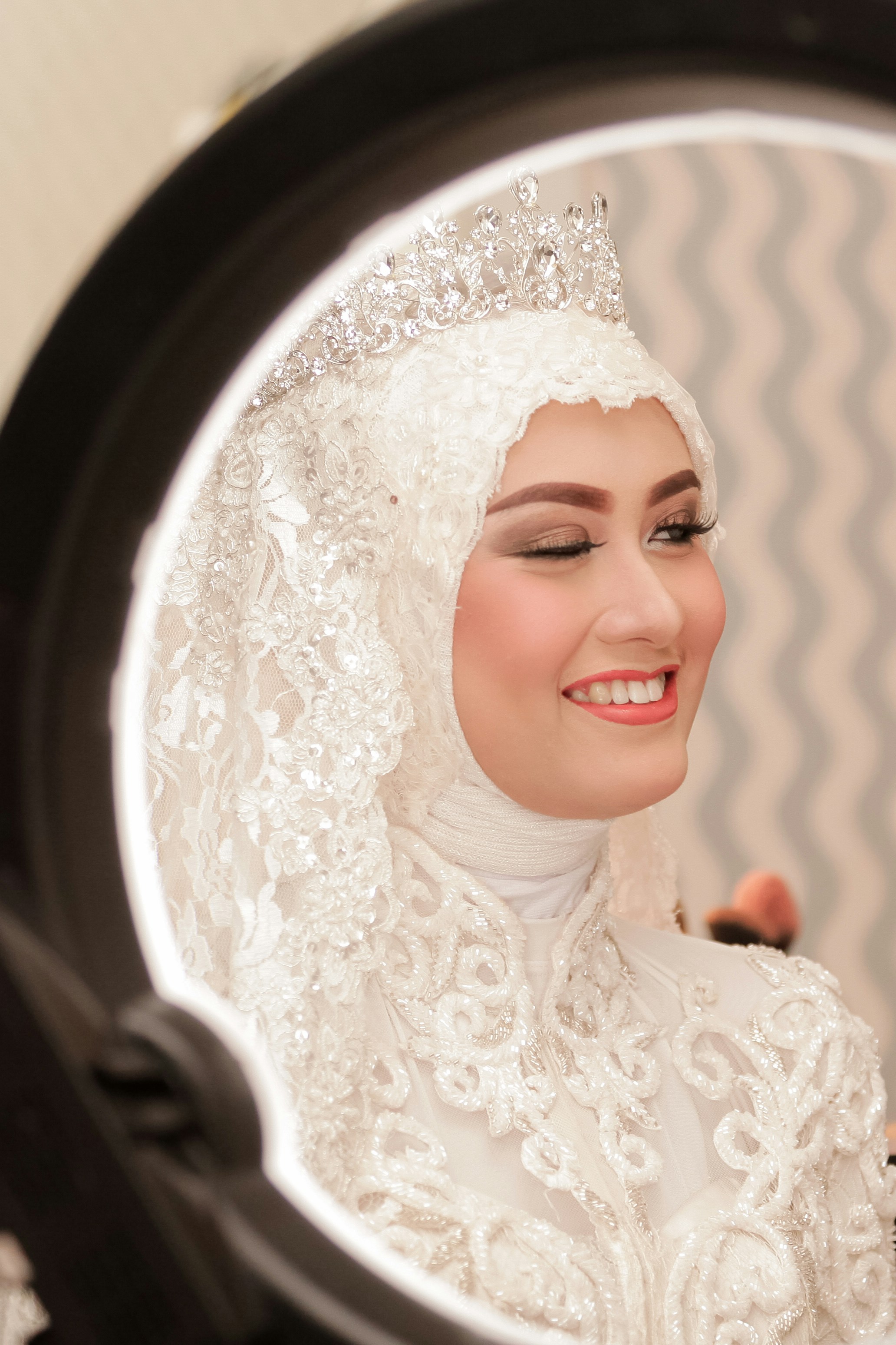 A smiling bride in a white lace hijab and sparkling tiara looking into a mirror