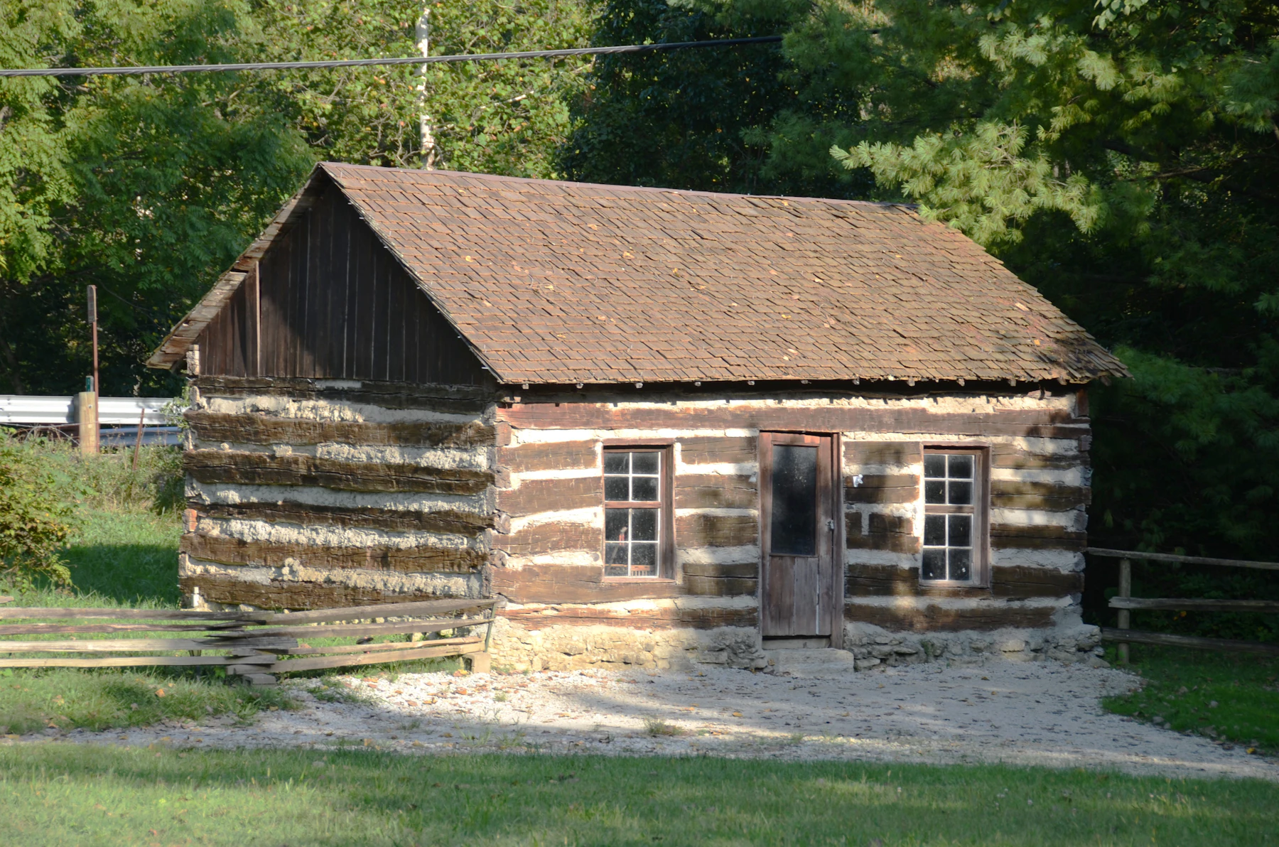 A one-story chinked log cabin with a door and two windows standing in a wooded clearing