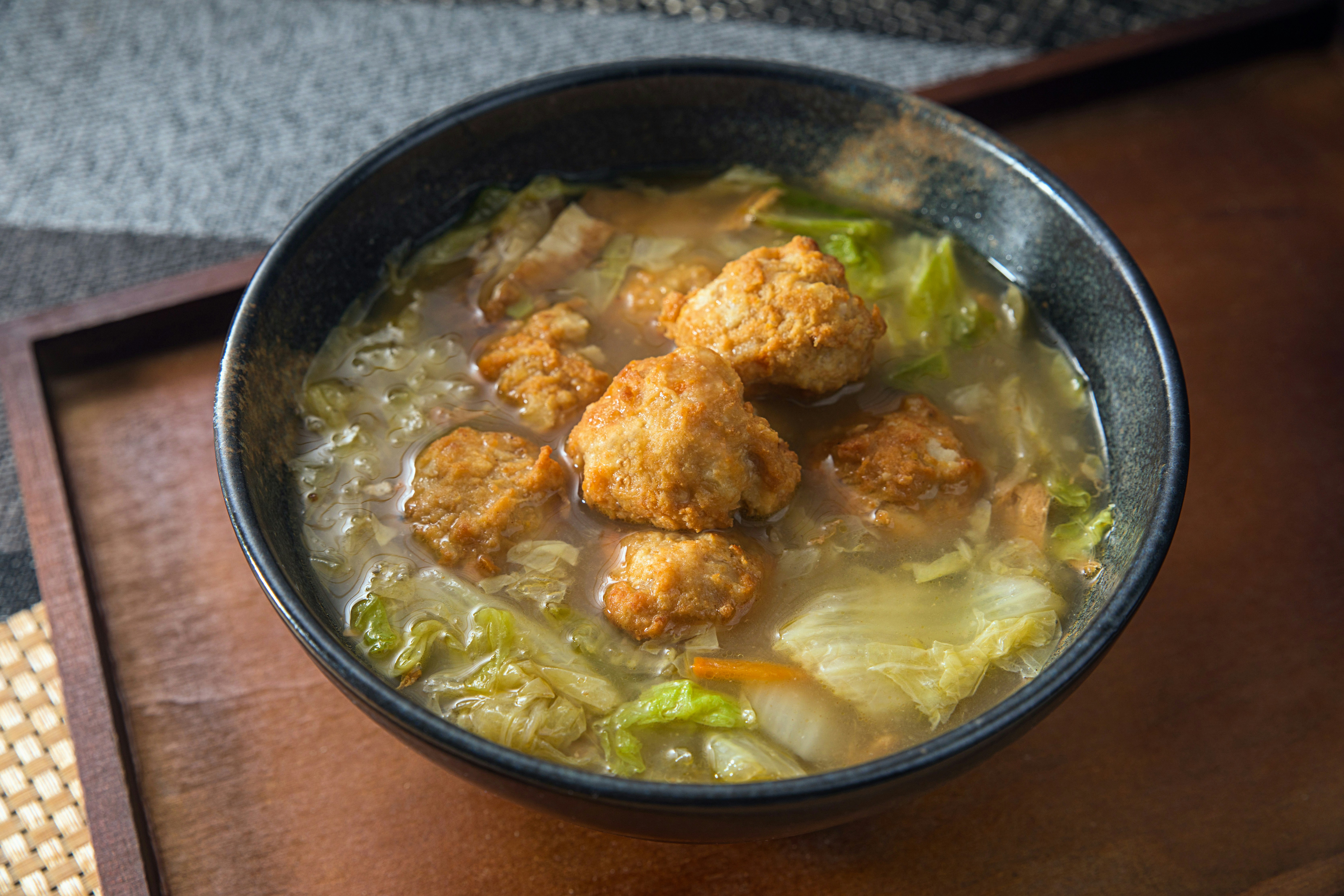 A bowl of soup with fried meatballs and cabbage on a wooden tray