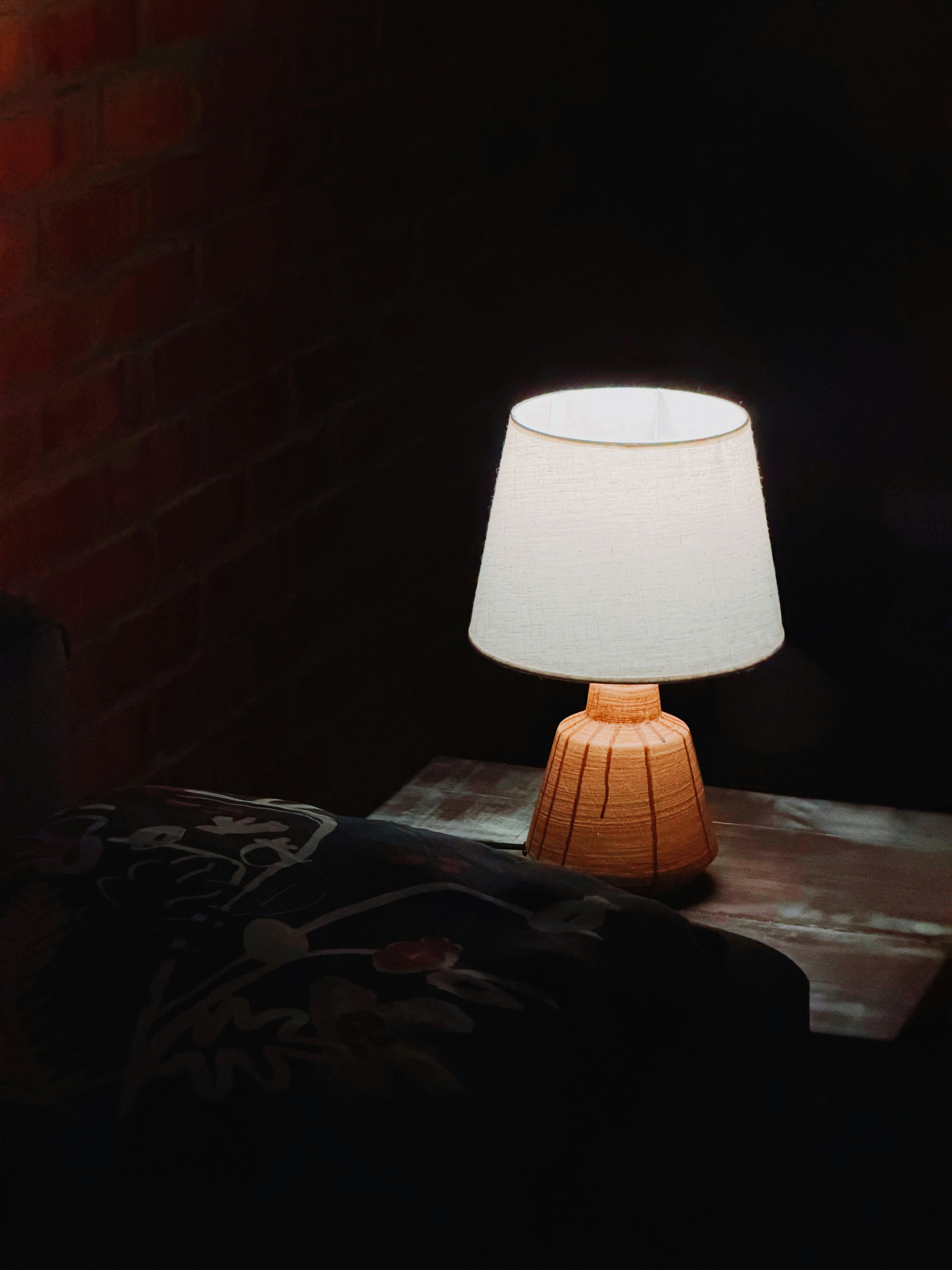 A lit table lamp with a textured base on a wooden surface near brick