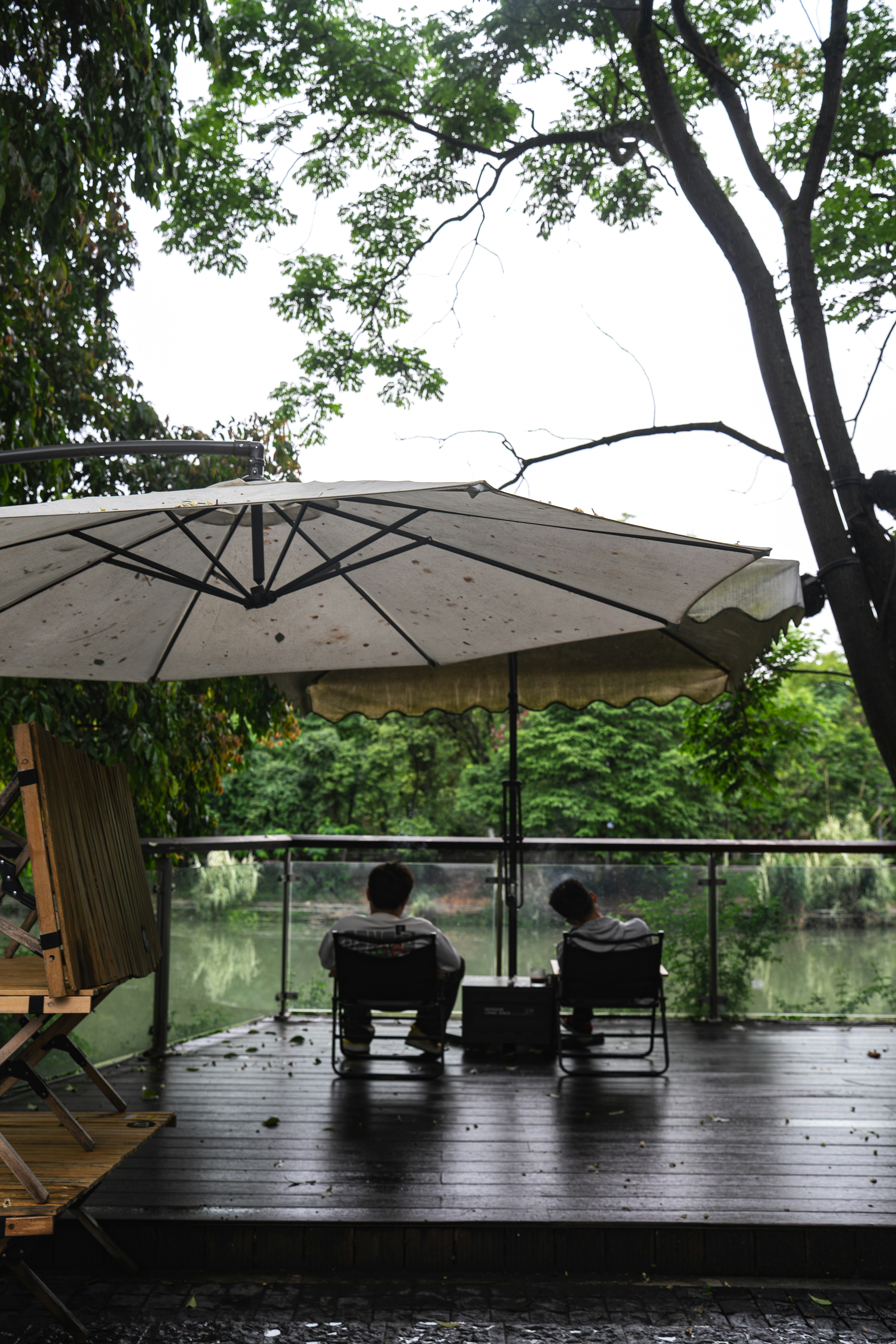 Two people in folding chairs under a large umbrella on a wooden deck