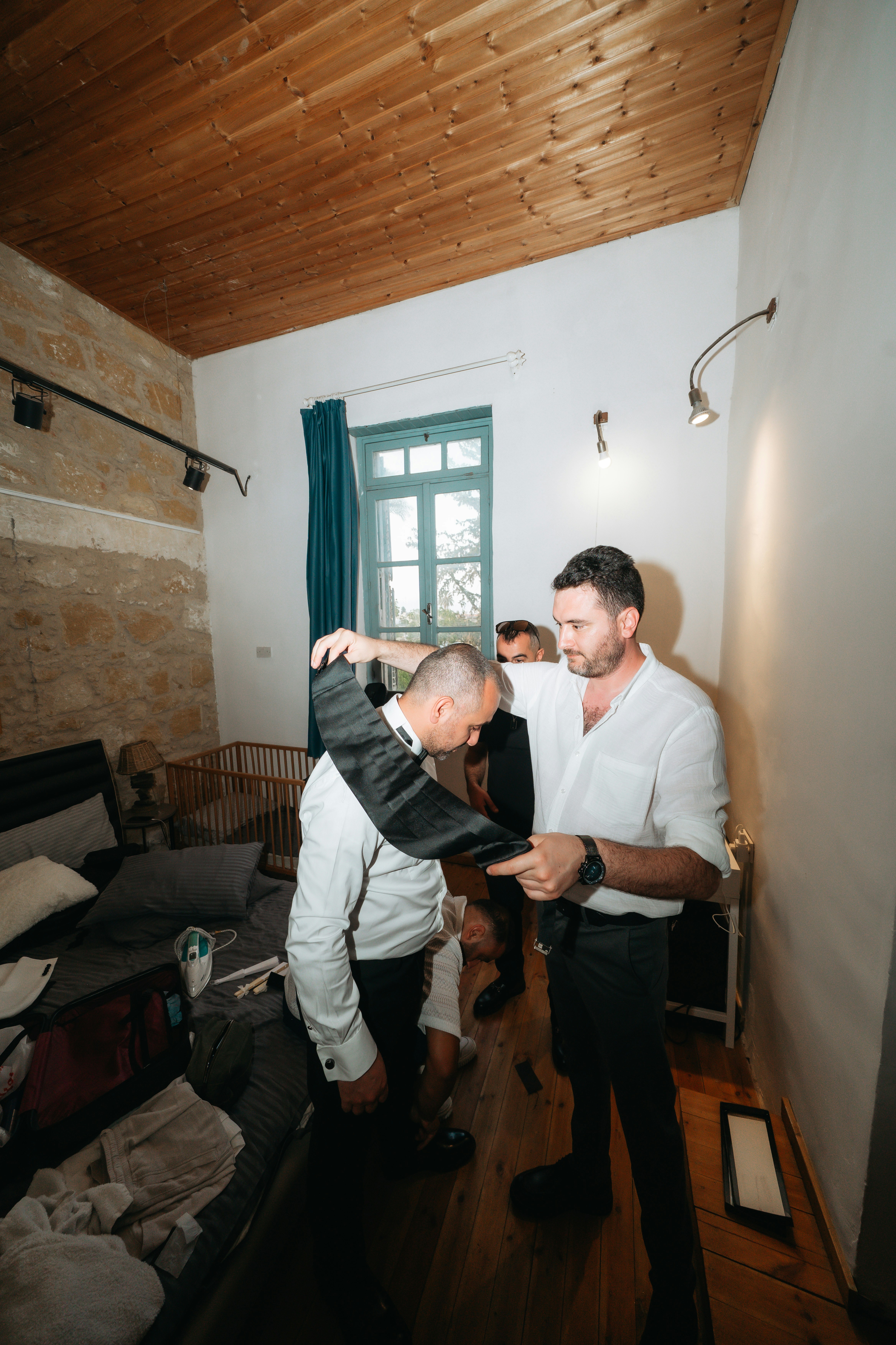 A man helping another man put on a black cummerbund in a bedroom