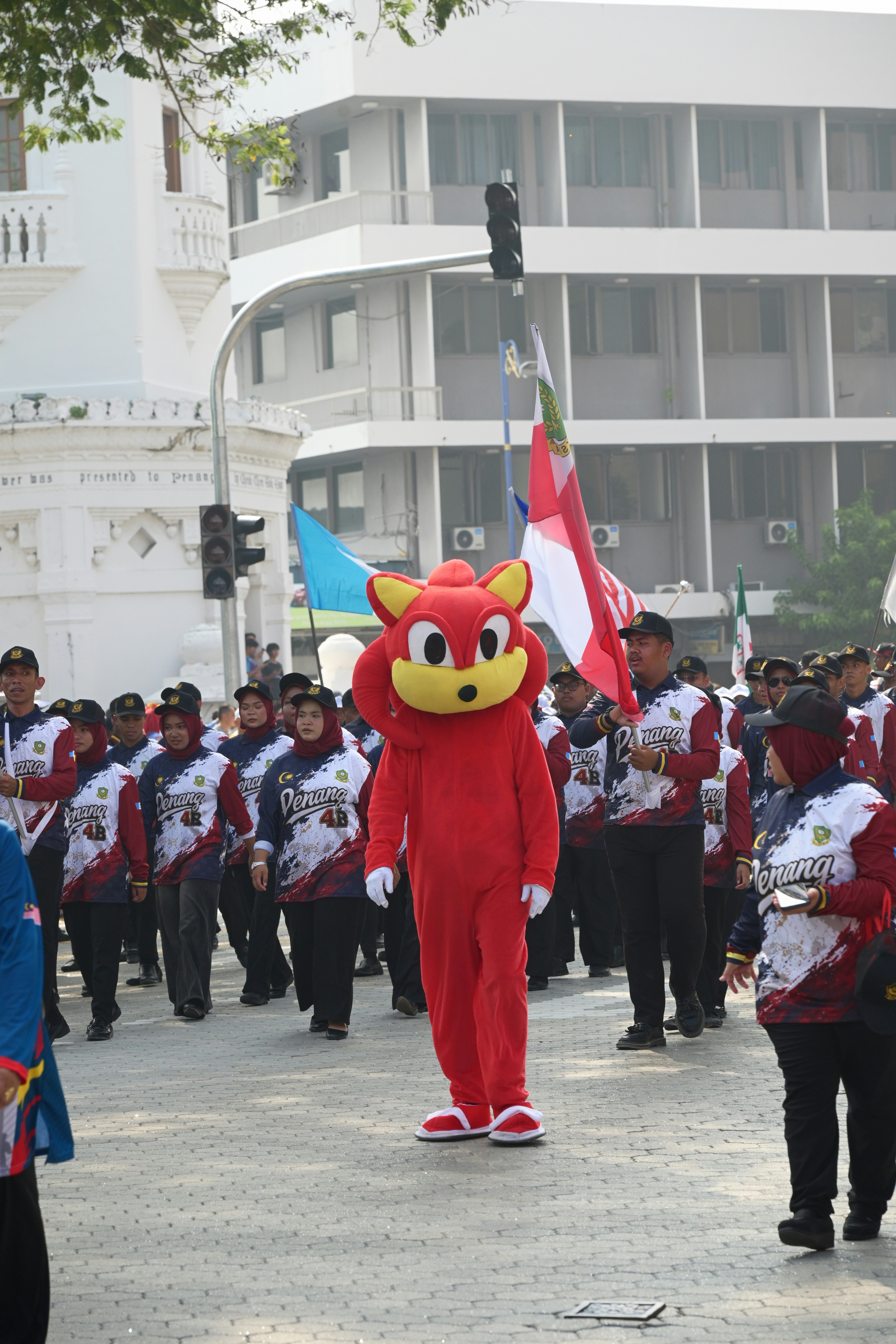 A red mascot walking in a parade with people in matching uniforms in Penang