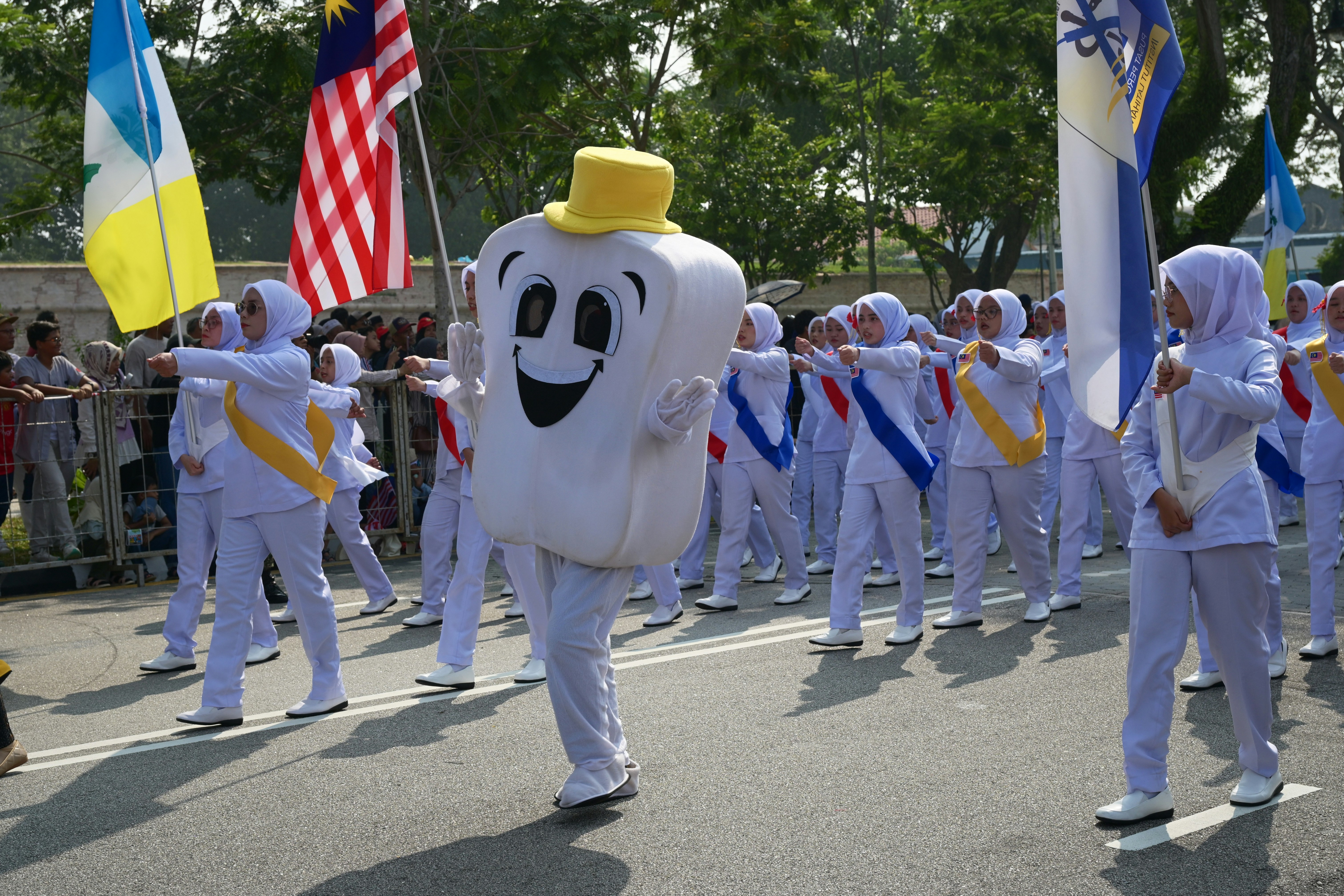 A tooth mascot in a yellow hat leading a marching group in white uniforms