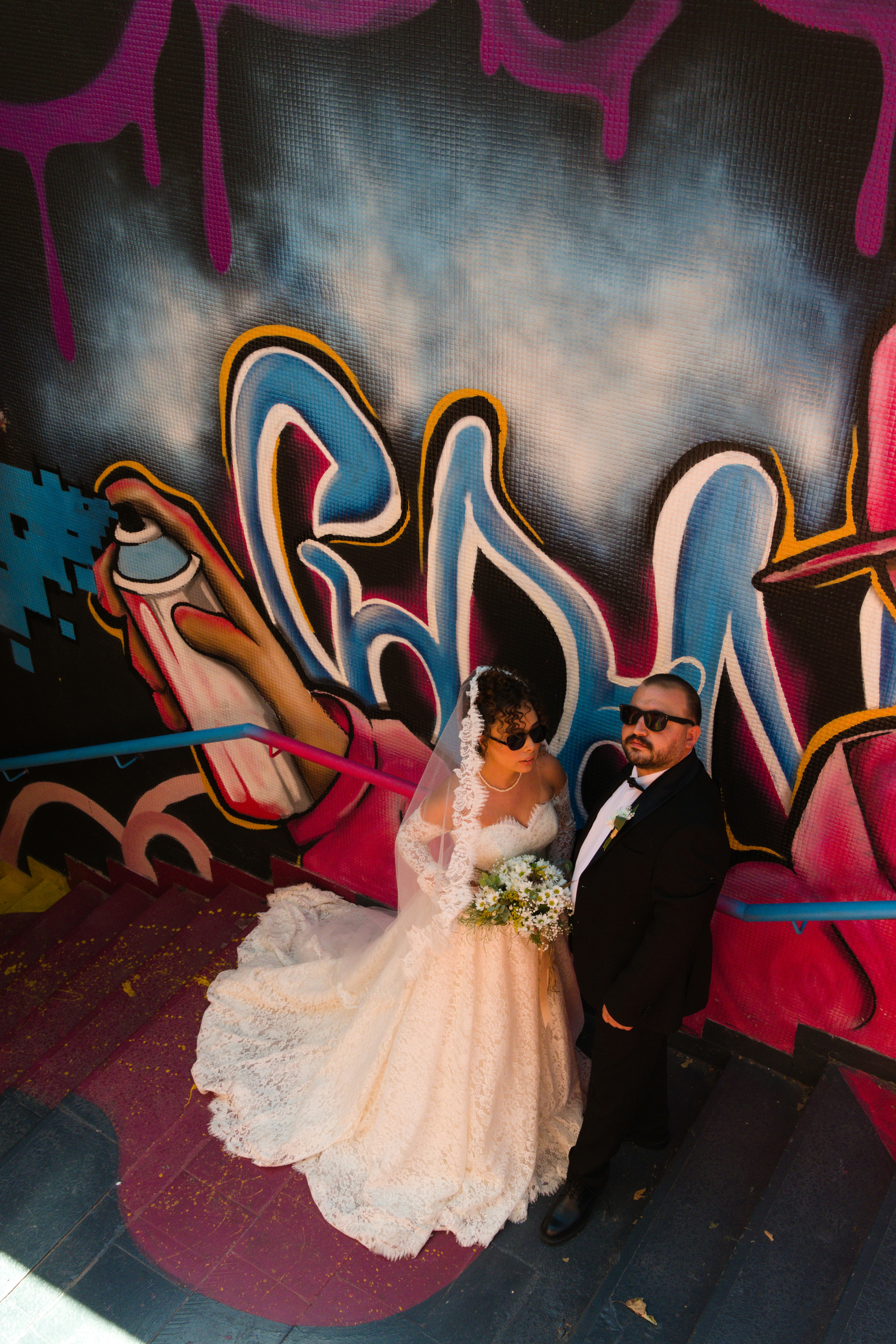 A bride and groom in sunglasses on stairs before a colorful graffiti wall