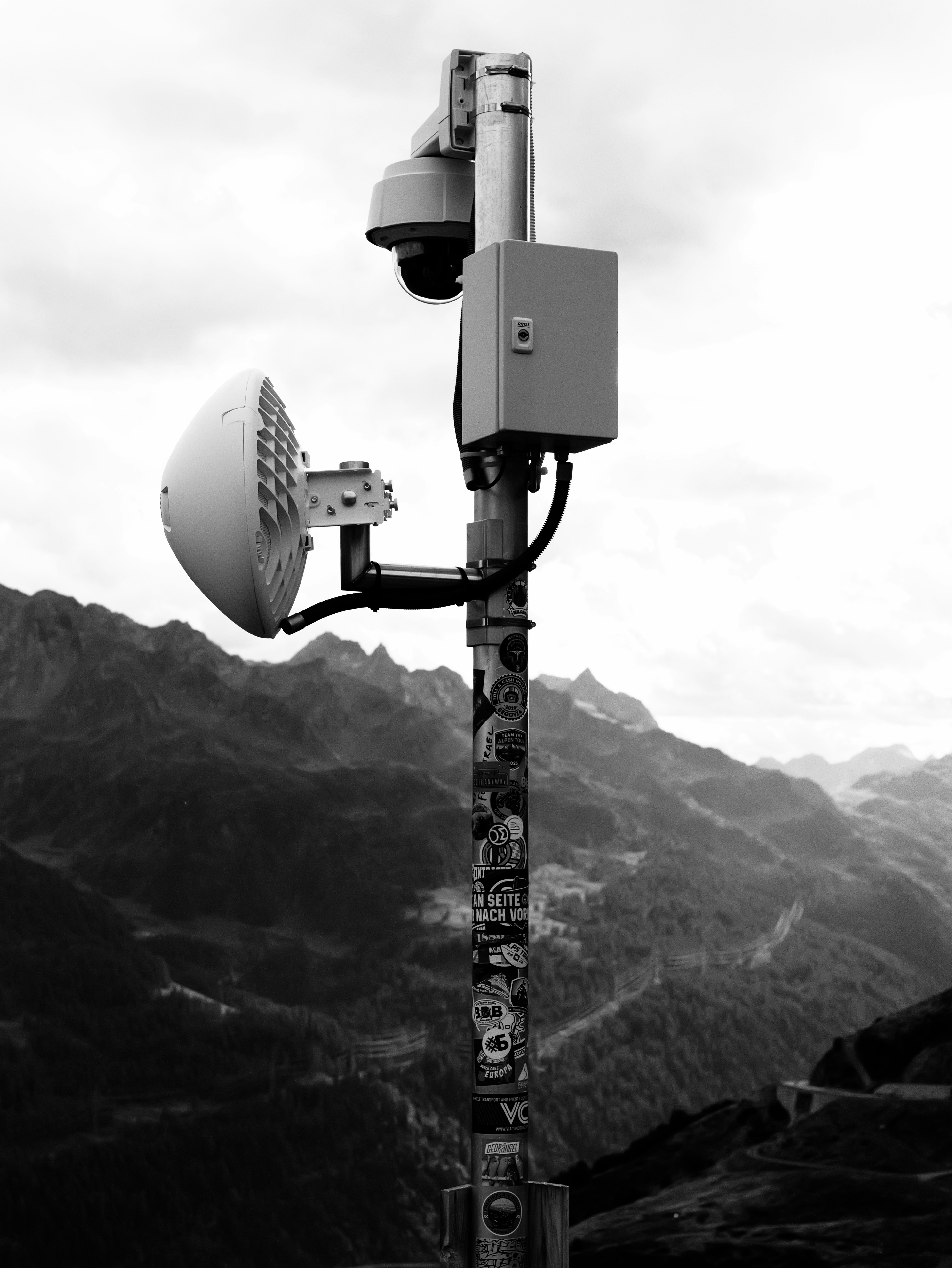 A surveillance camera and microwave antenna on a sticker-covered pole in the mountains