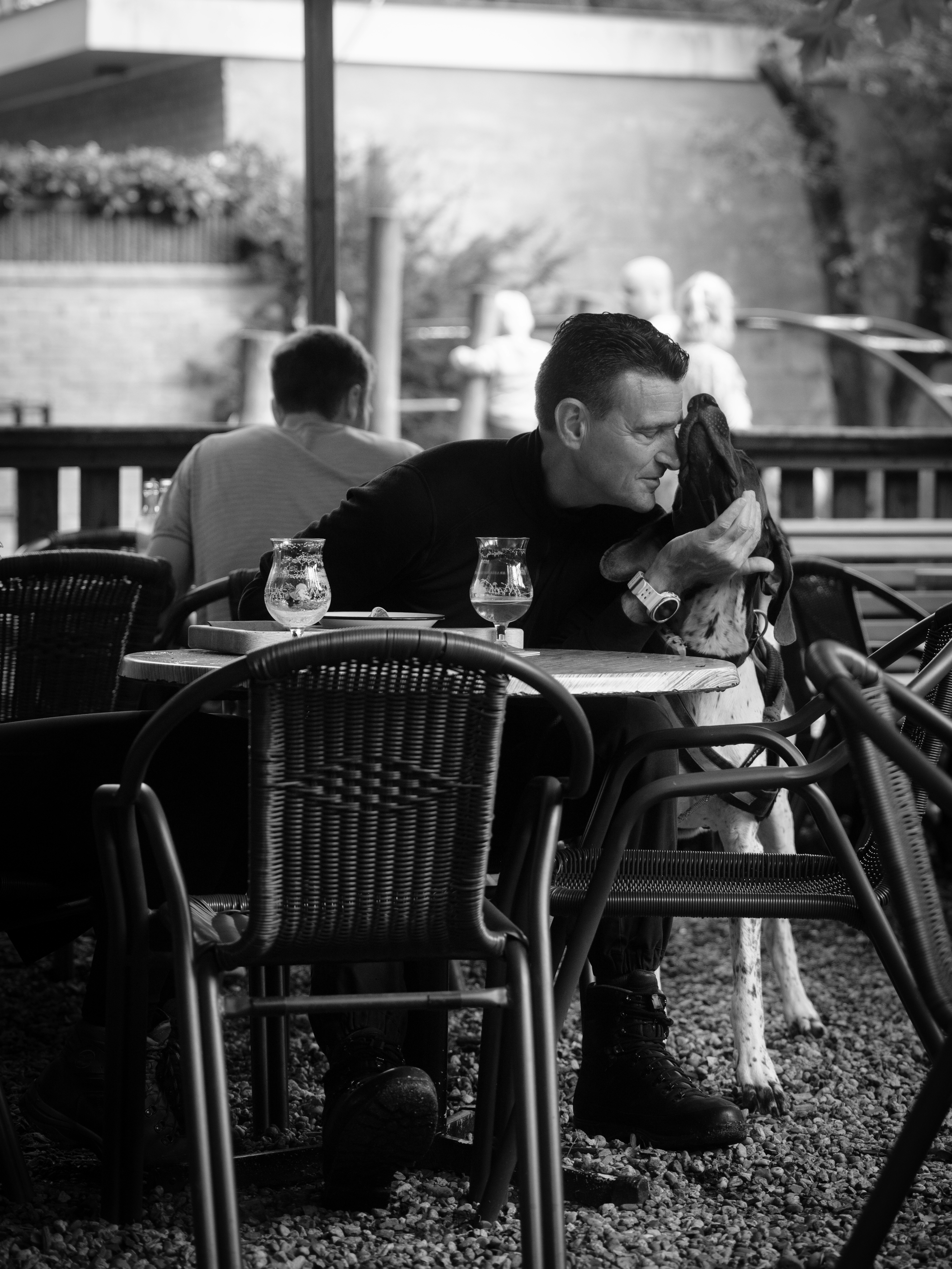 A man in a black shirt nuzzling a spotted dog at an outdoor cafe
