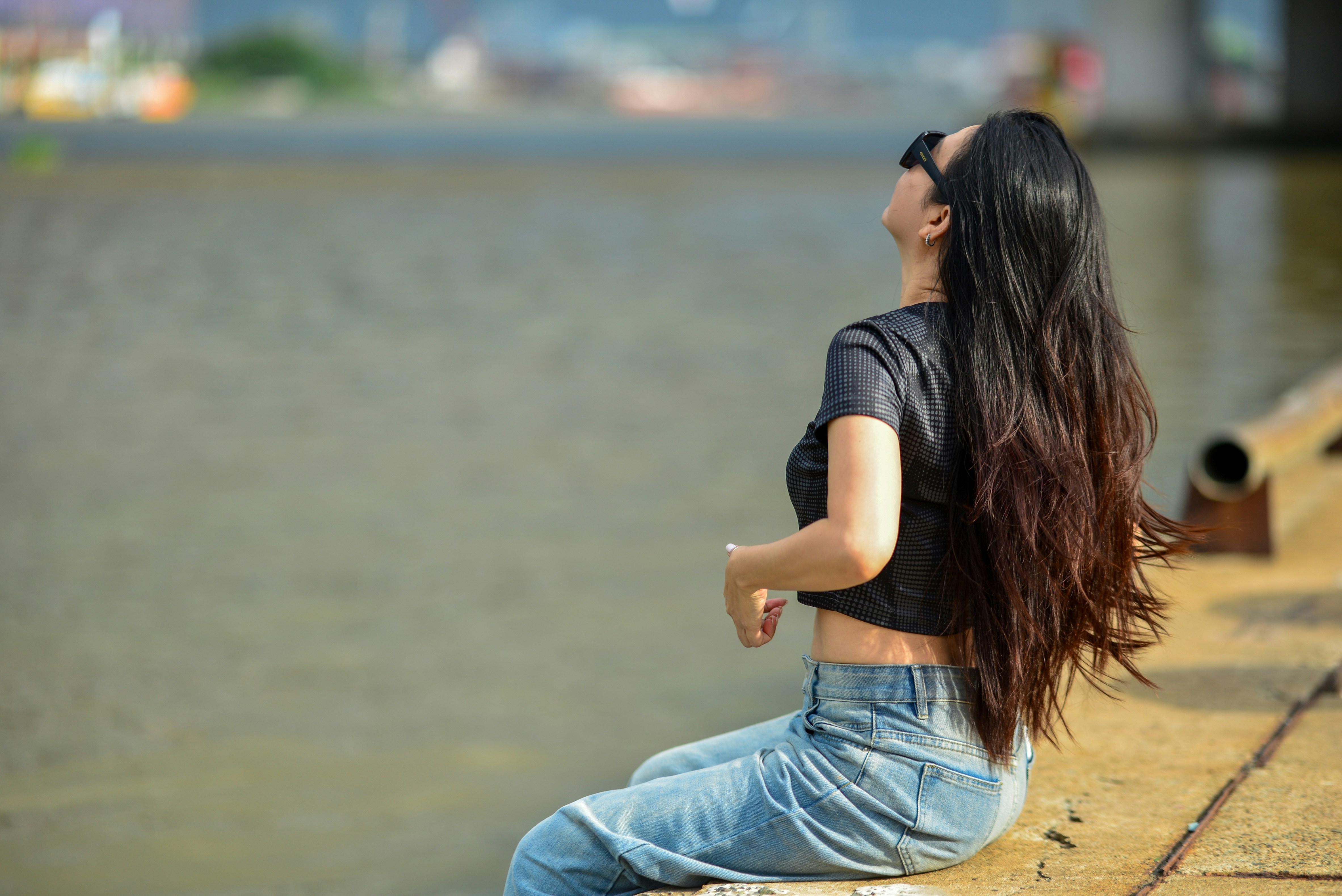 A woman with long dark hair wearing sunglasses sitting by a river