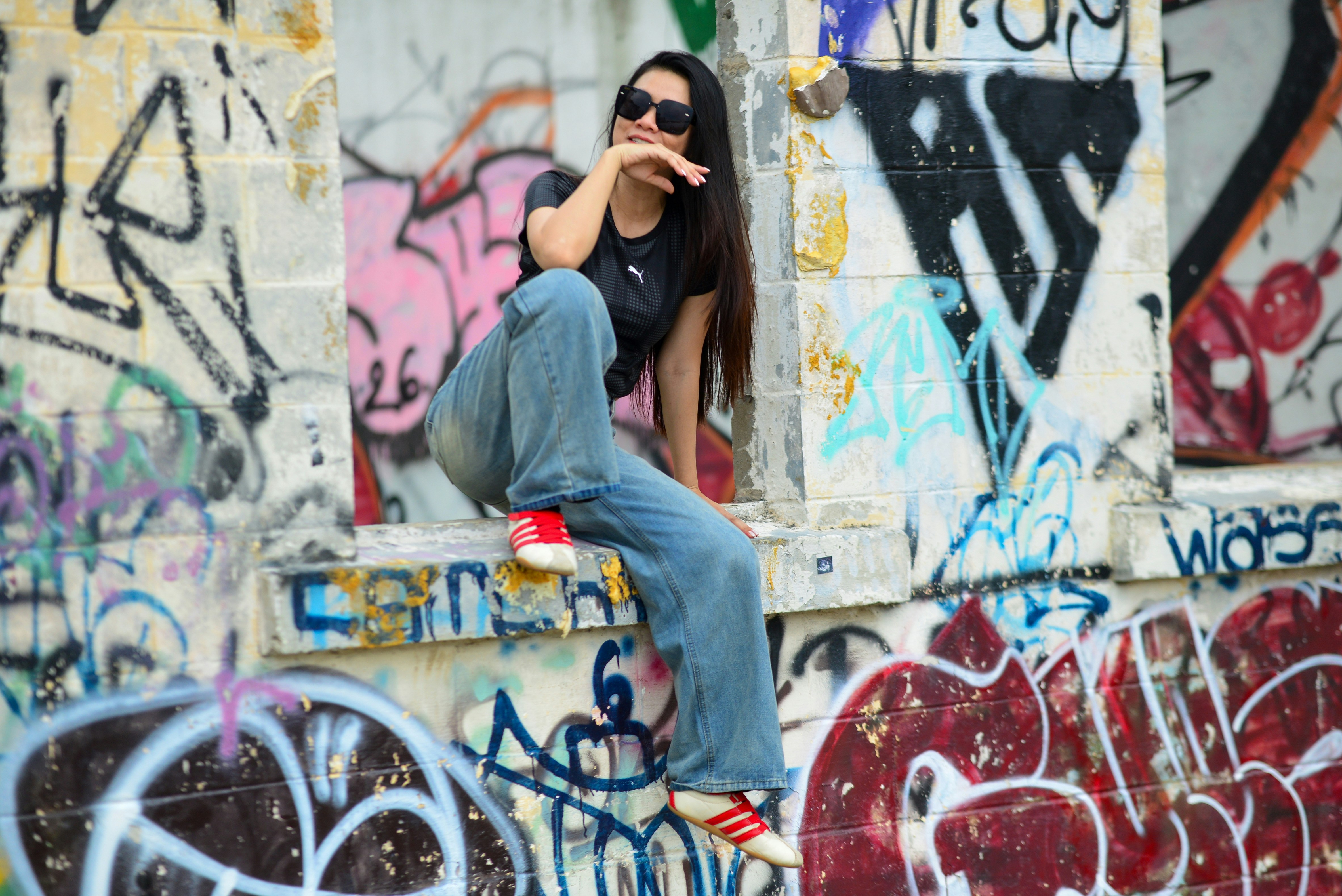 A woman in sunglasses and jeans sitting on a graffiti covered concrete wall