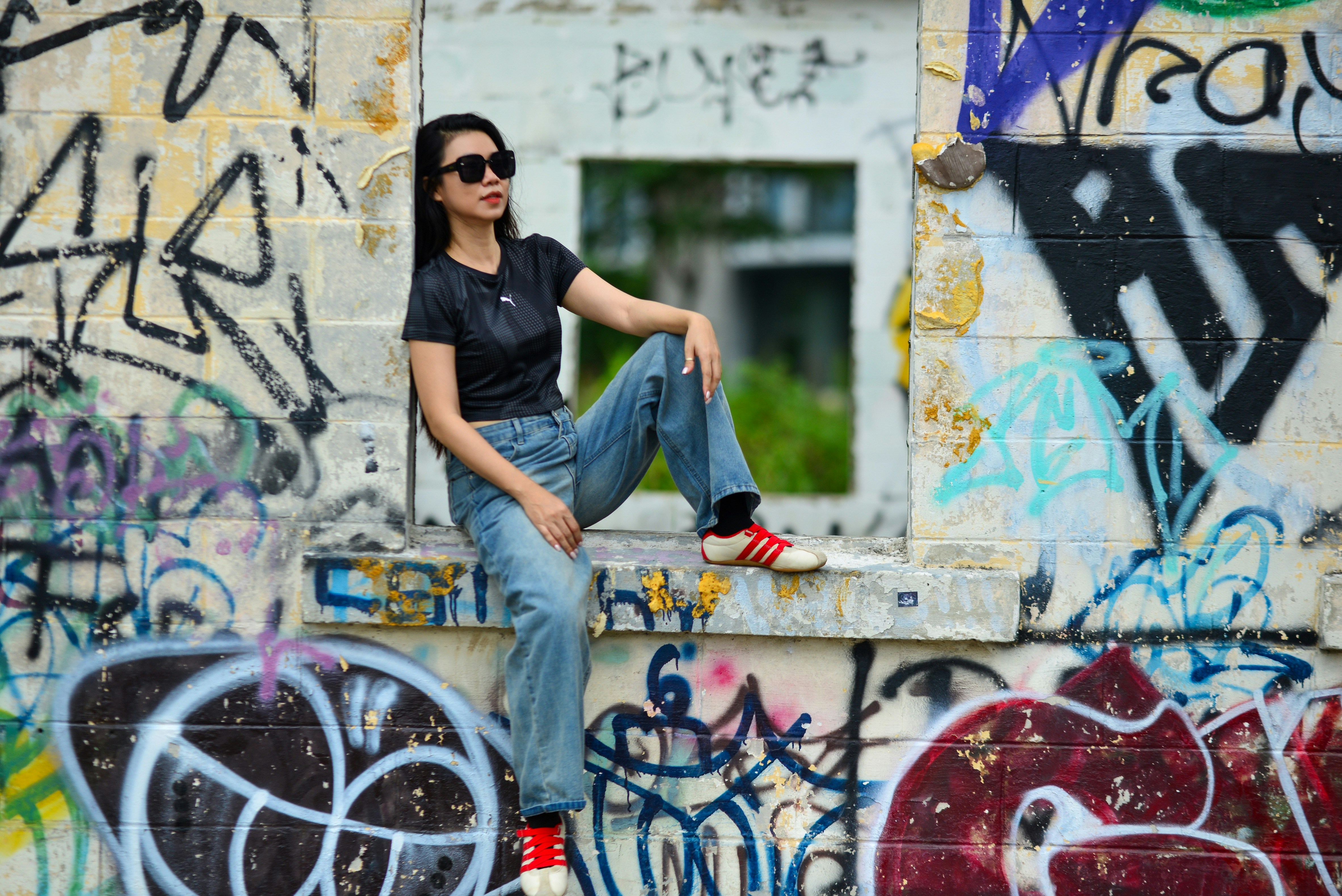 A woman in sunglasses and a black t-shirt sitting in a graffiti-covered window frame