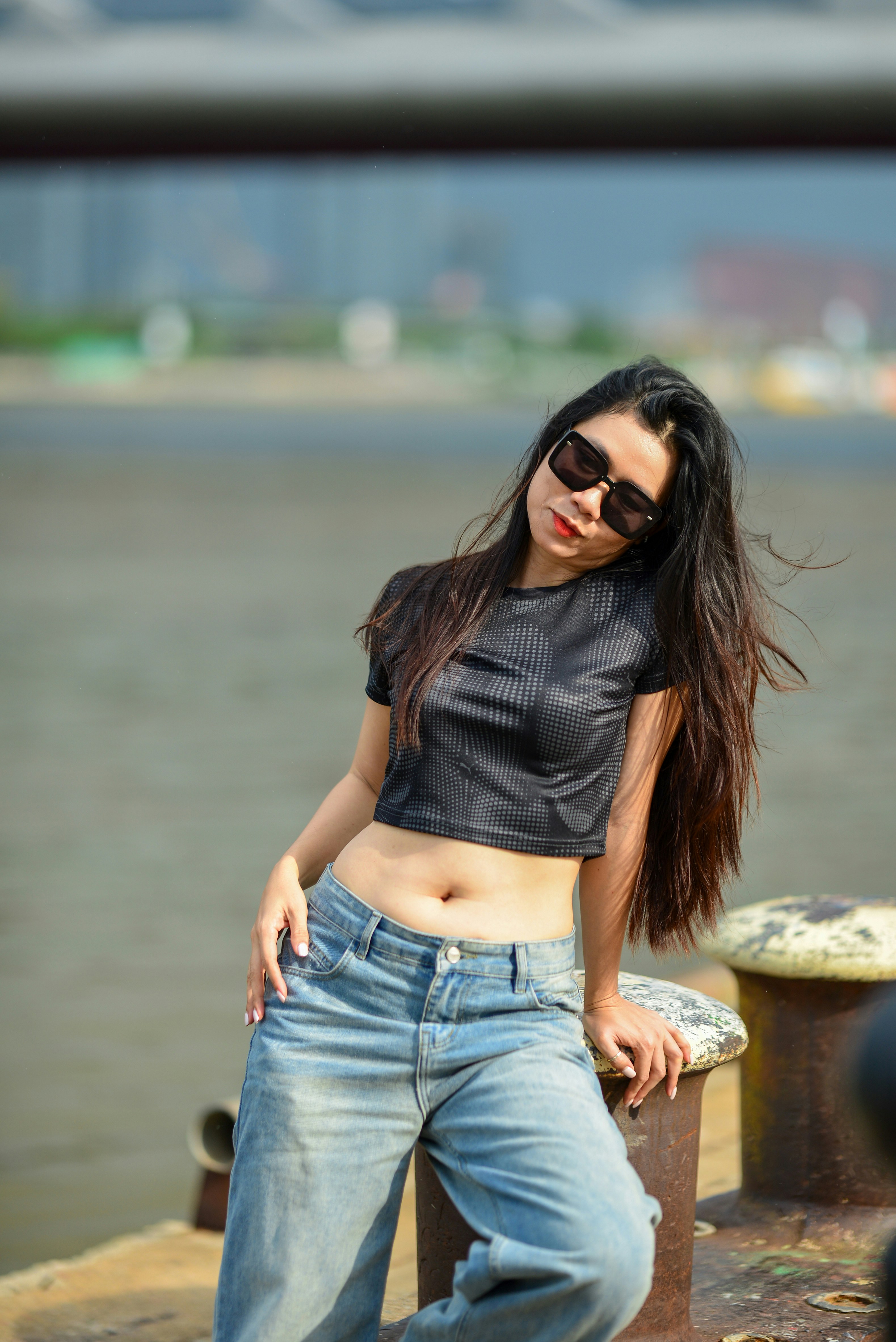 A woman with long dark hair wearing sunglasses and a black crop top by water