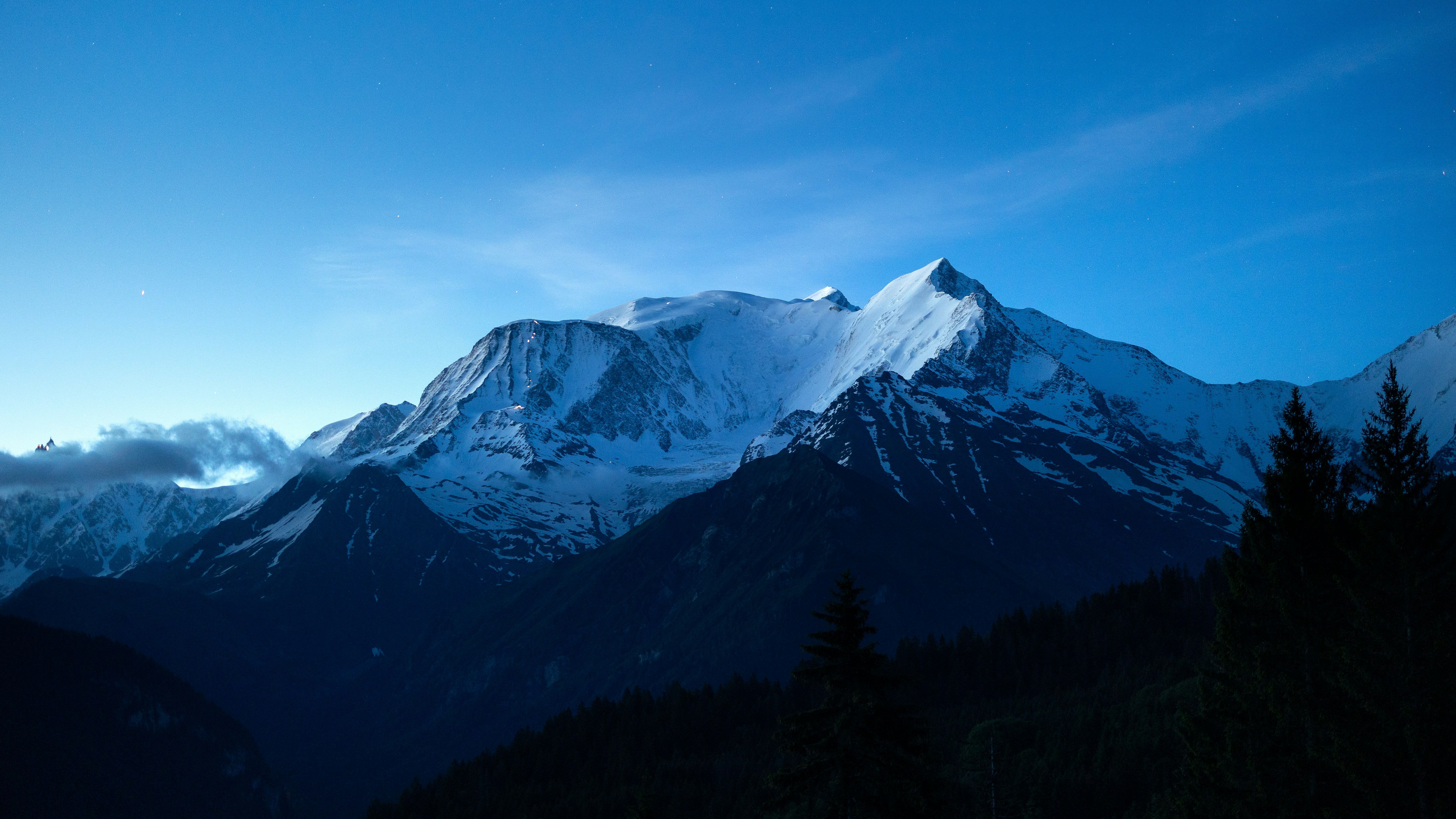Snow-covered Mont Blanc peak under a clear blue night sky with dark pine trees