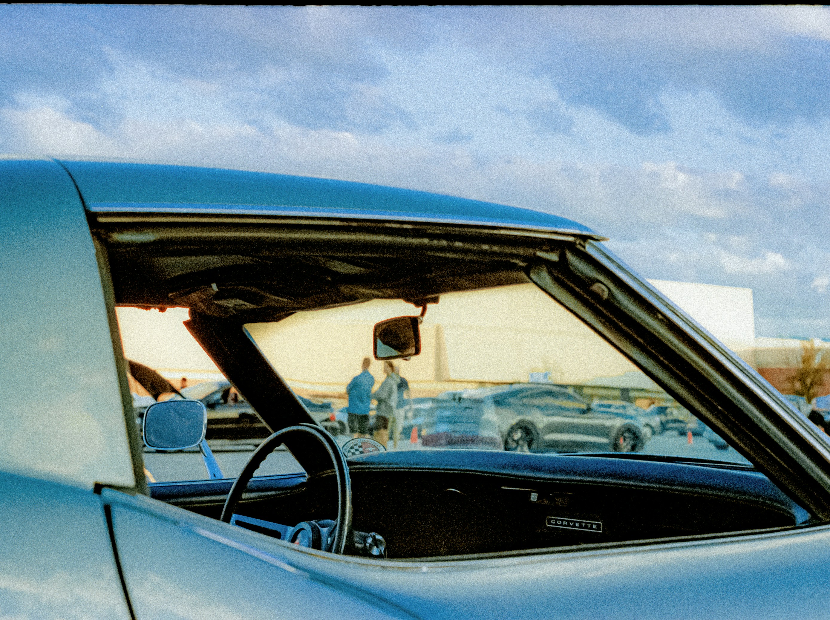 Interior view of a vintage Corvette looking out toward a parking lot gathering