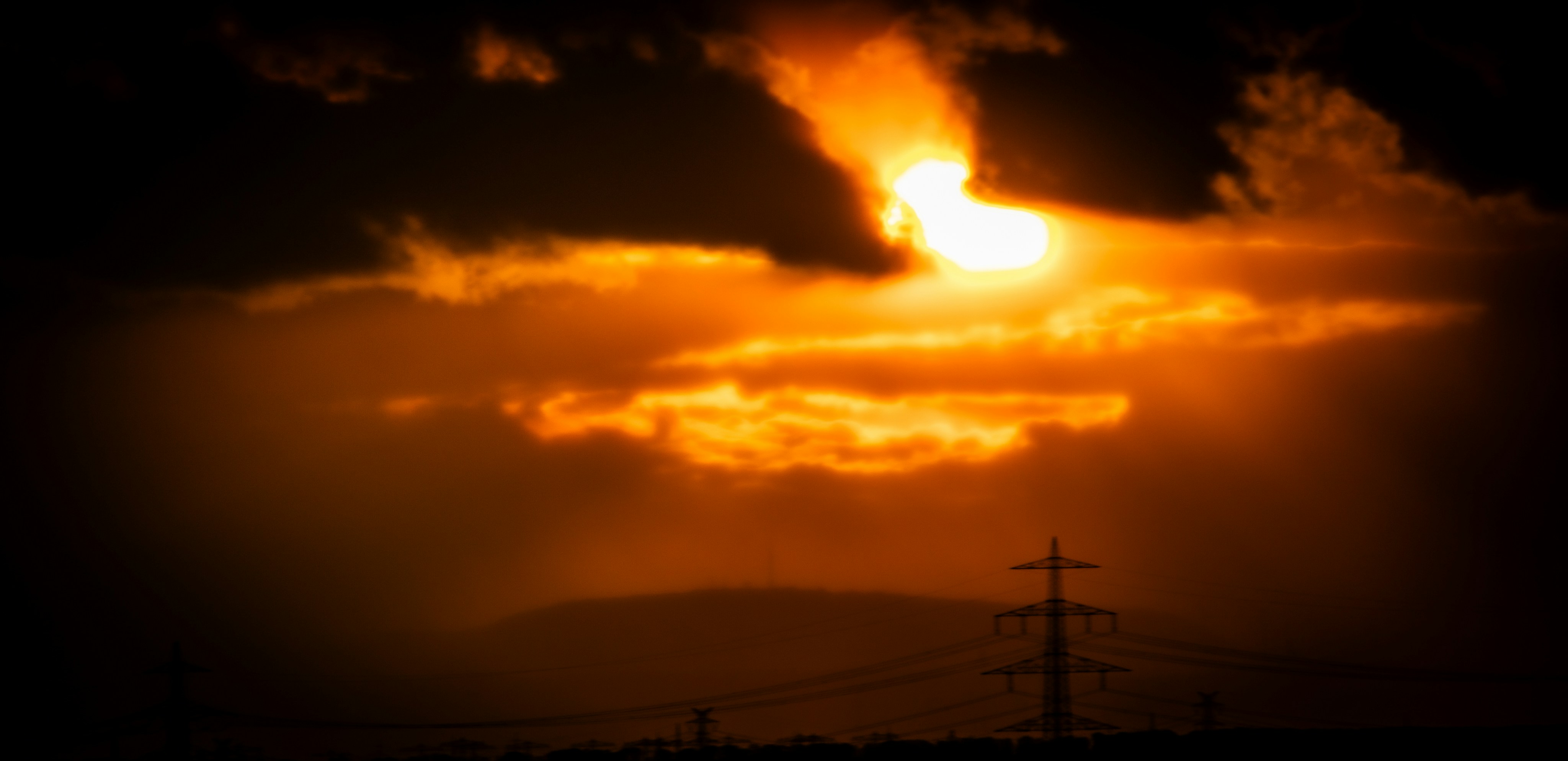 Low sun glowing behind dark clouds above a silhouette of electricity pylons