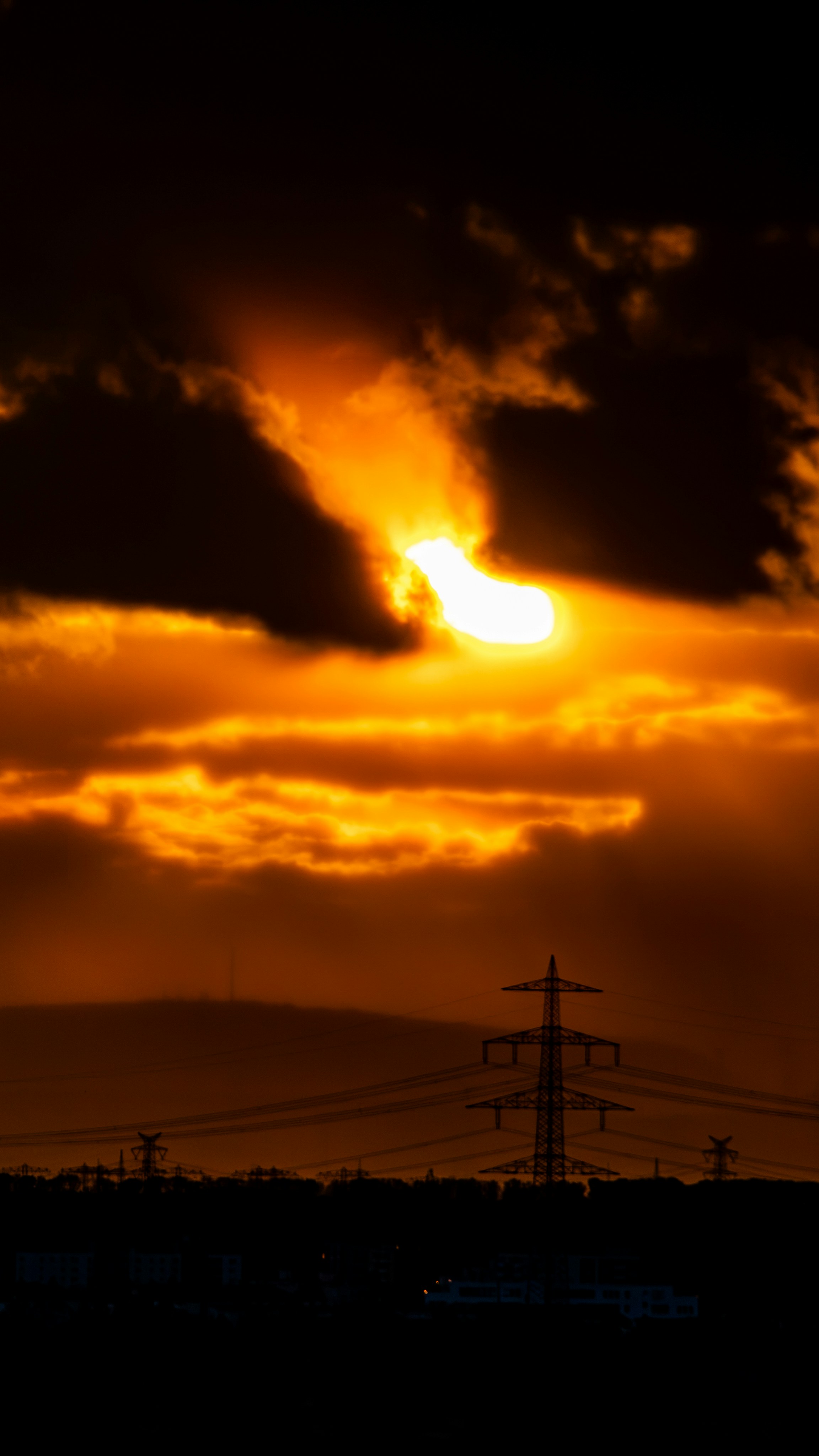A bright sun glowing behind dark clouds above a silhouette of power pylons