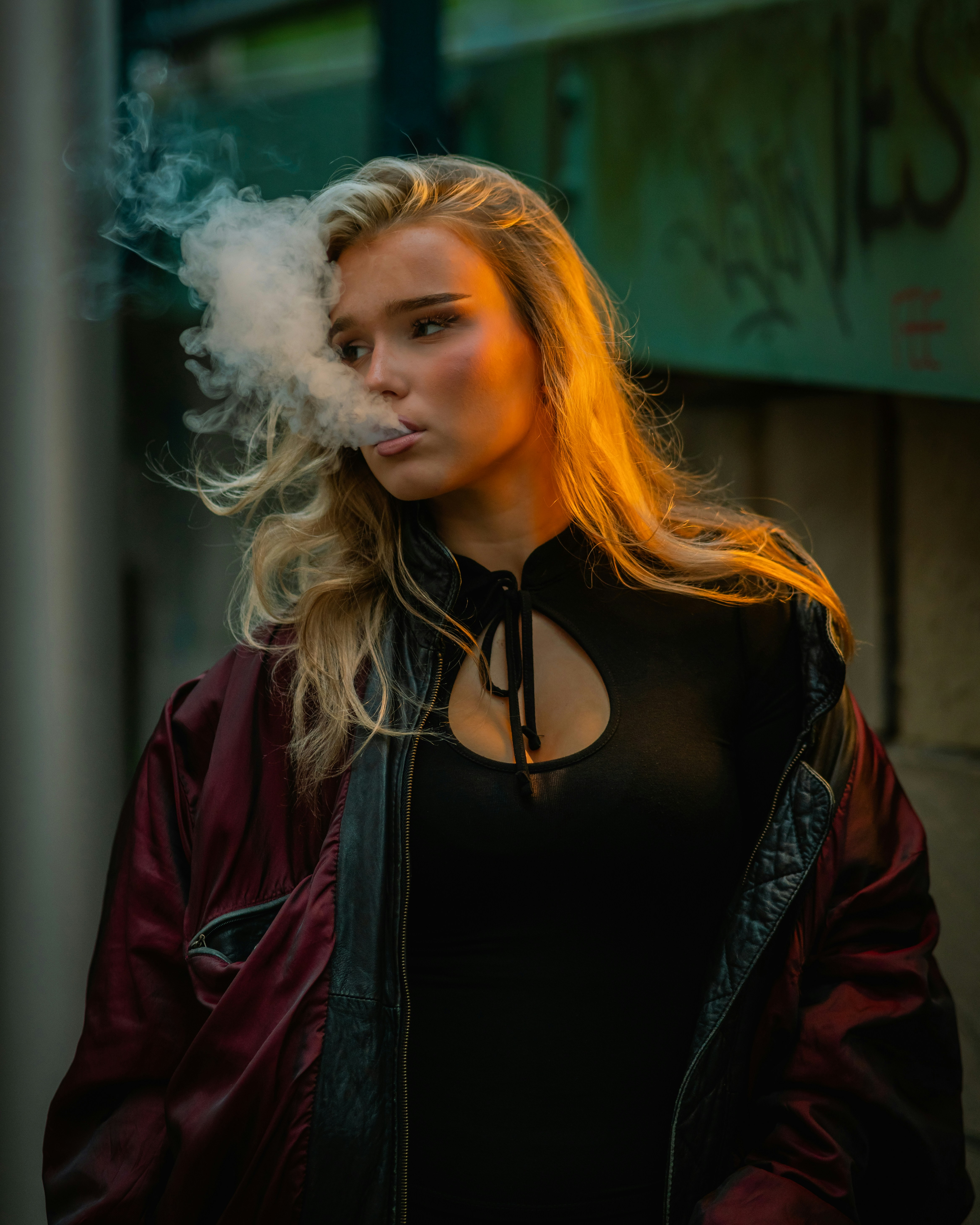 A woman in a dark jacket exhaling a cloud of smoke in golden light
