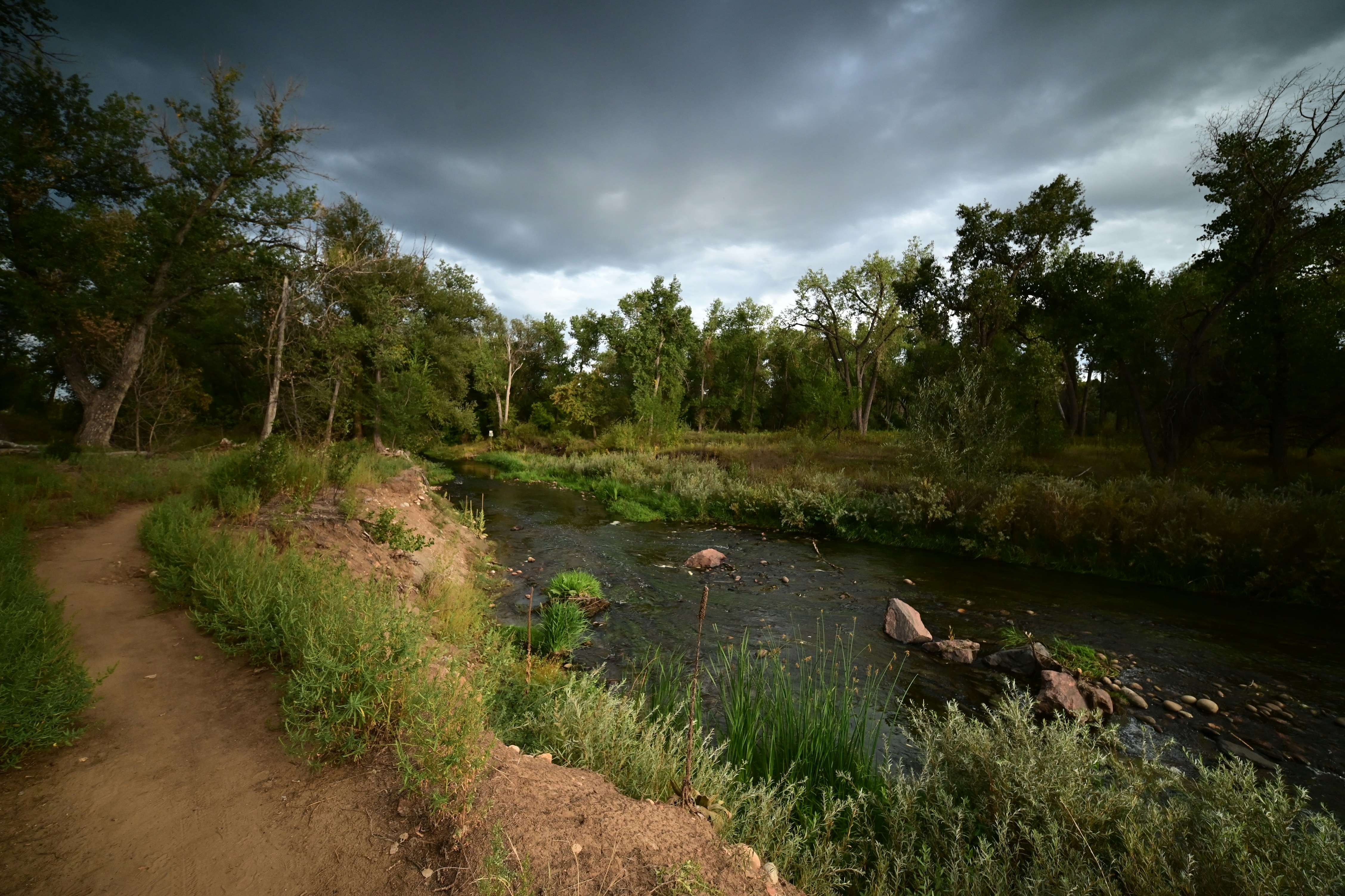 Dirt trail beside a shallow creek under a cloudy sky