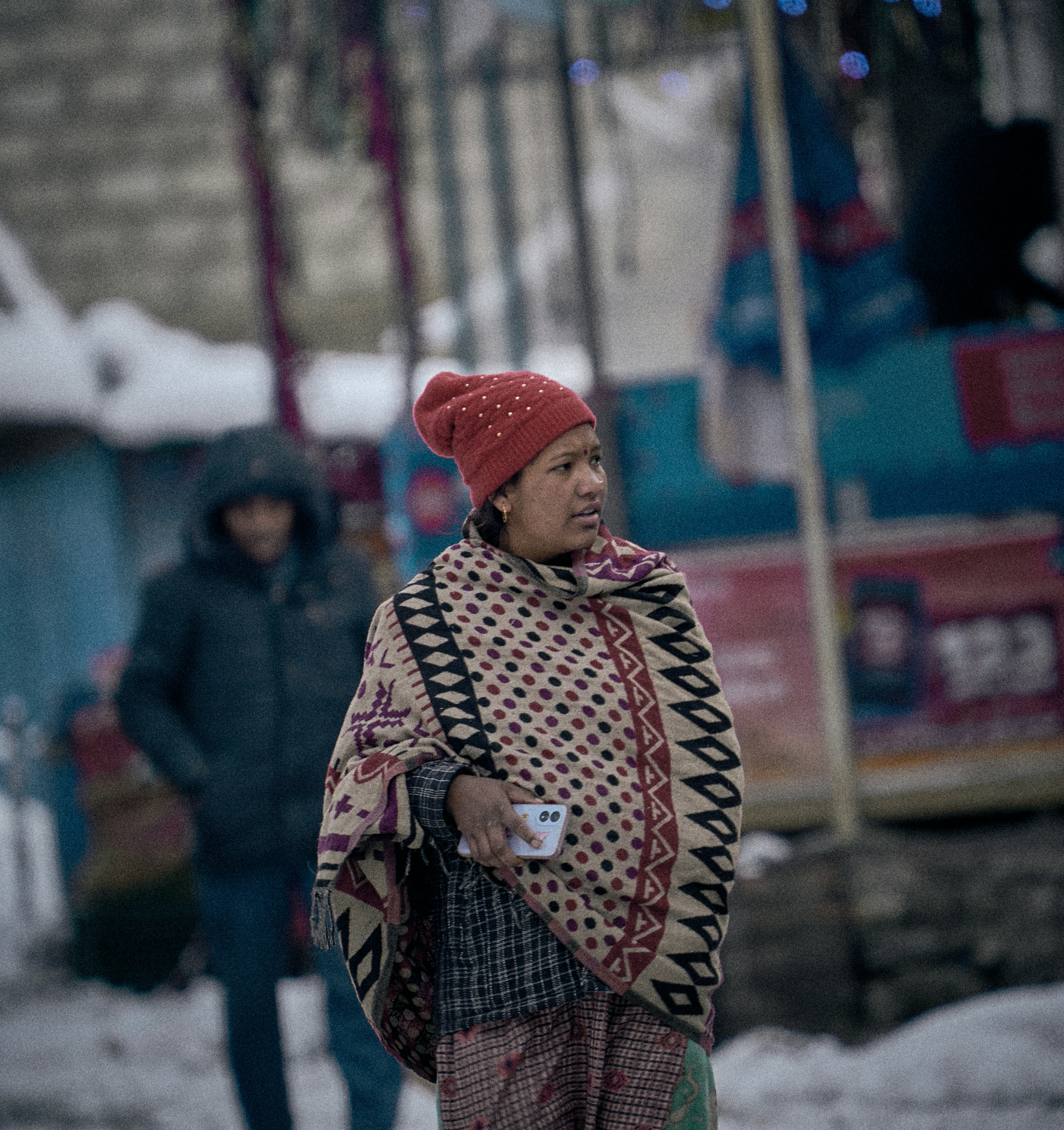 A woman in a red beanie and patterned shawl holding a smartphone outdoors