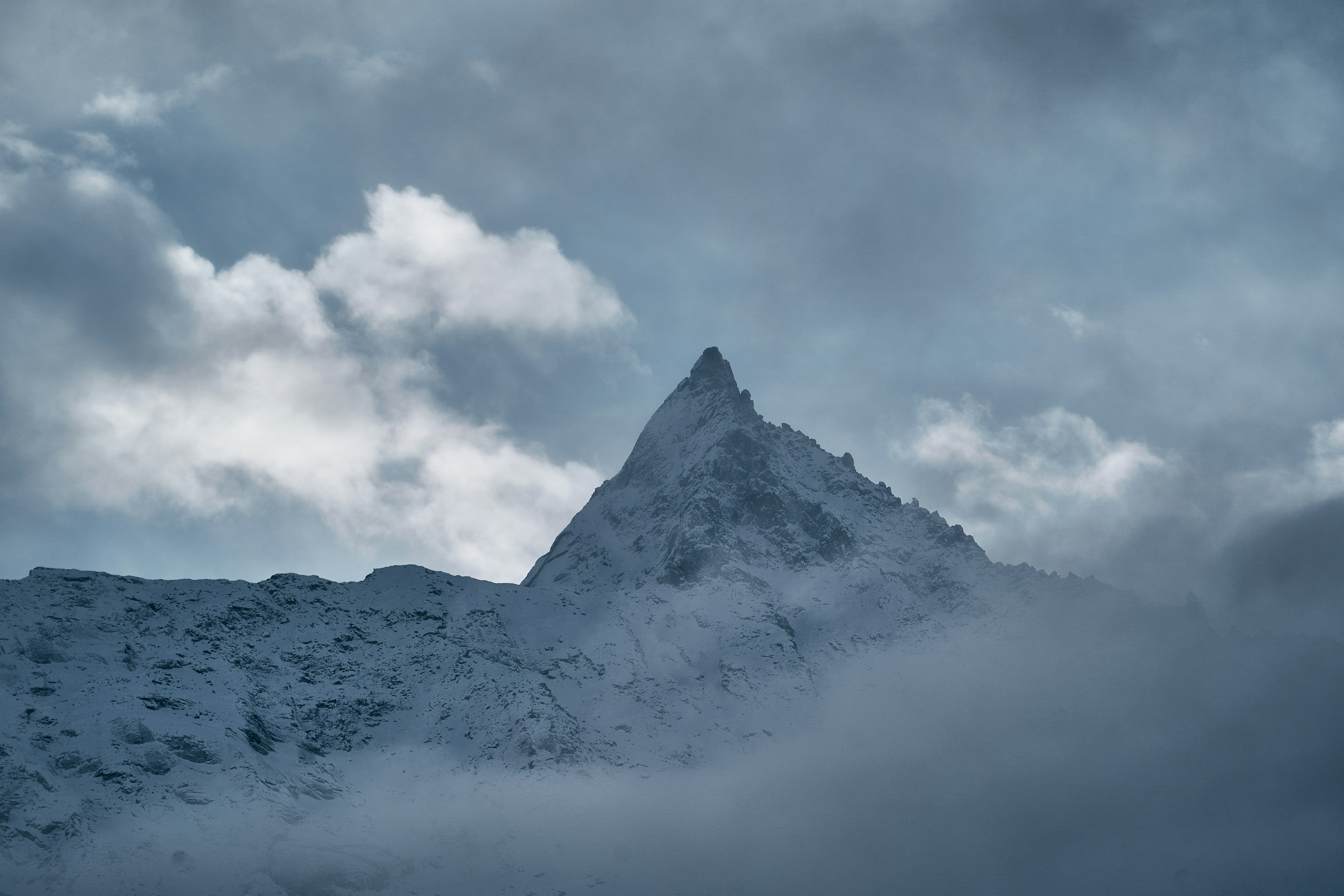 A sharp snow-covered mountain peak rising through thick grey clouds