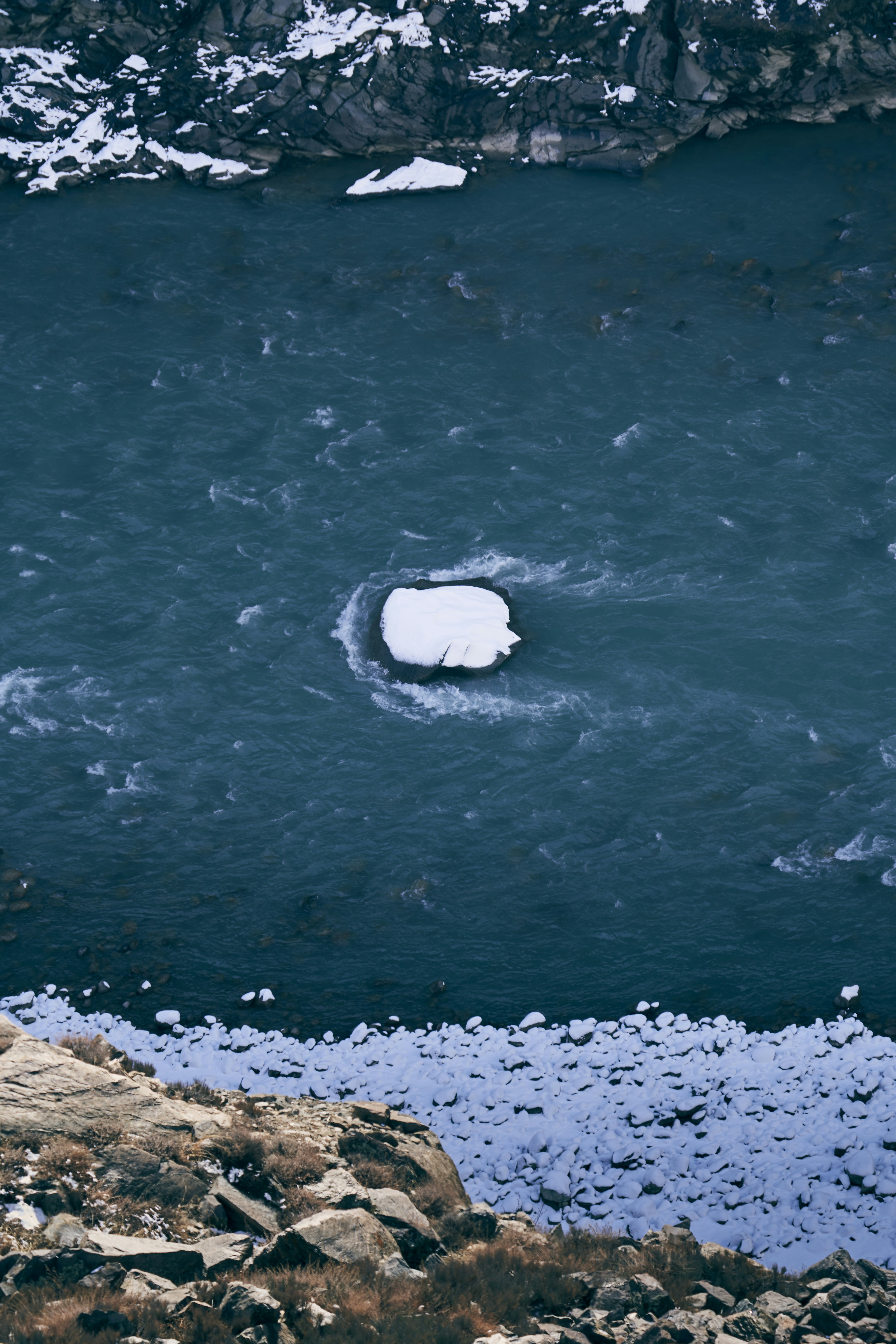 A snow-covered rock in the middle of a dark blue flowing river