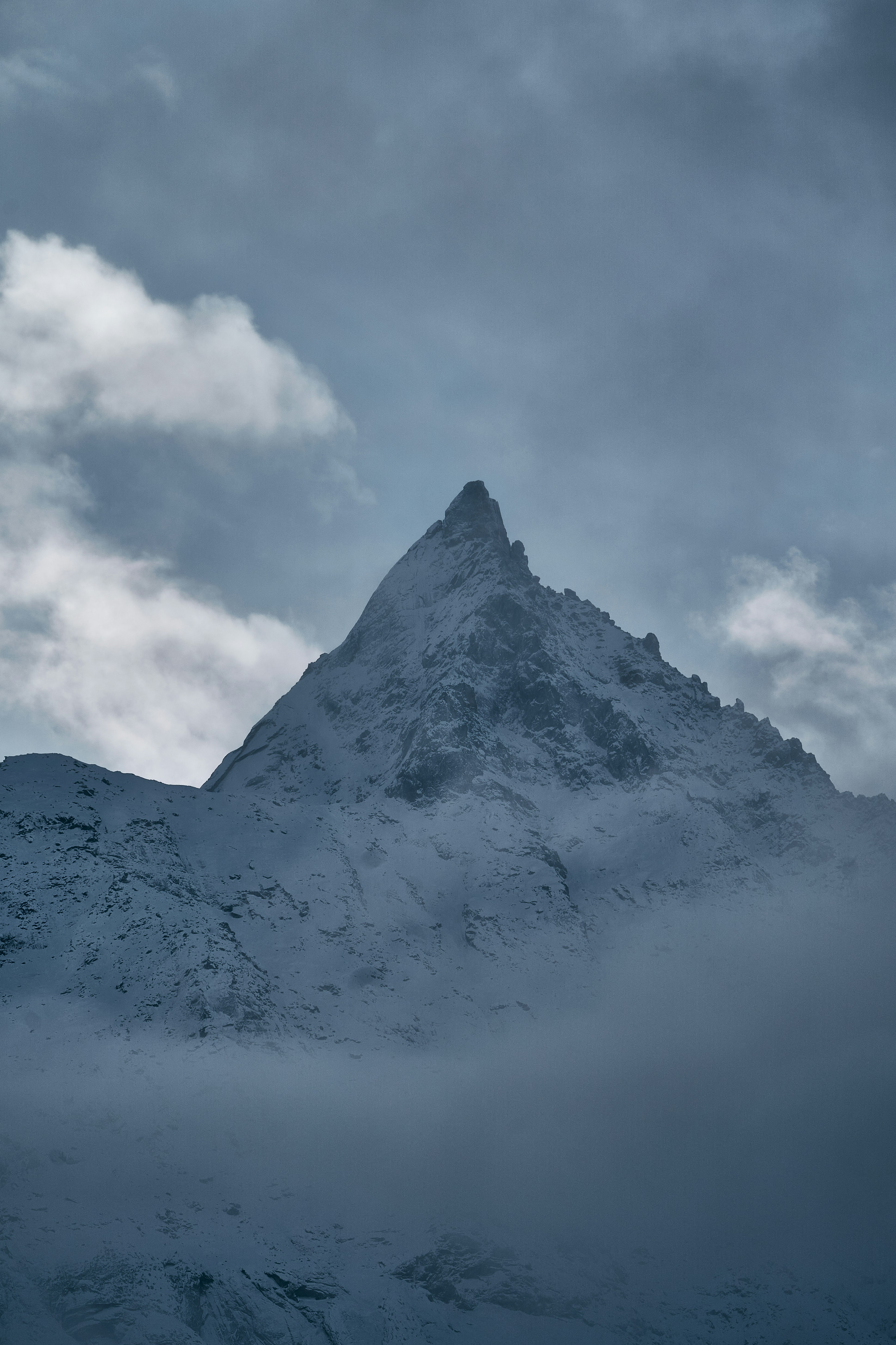 A snow-covered mountain peak rising through low clouds under a grey sky