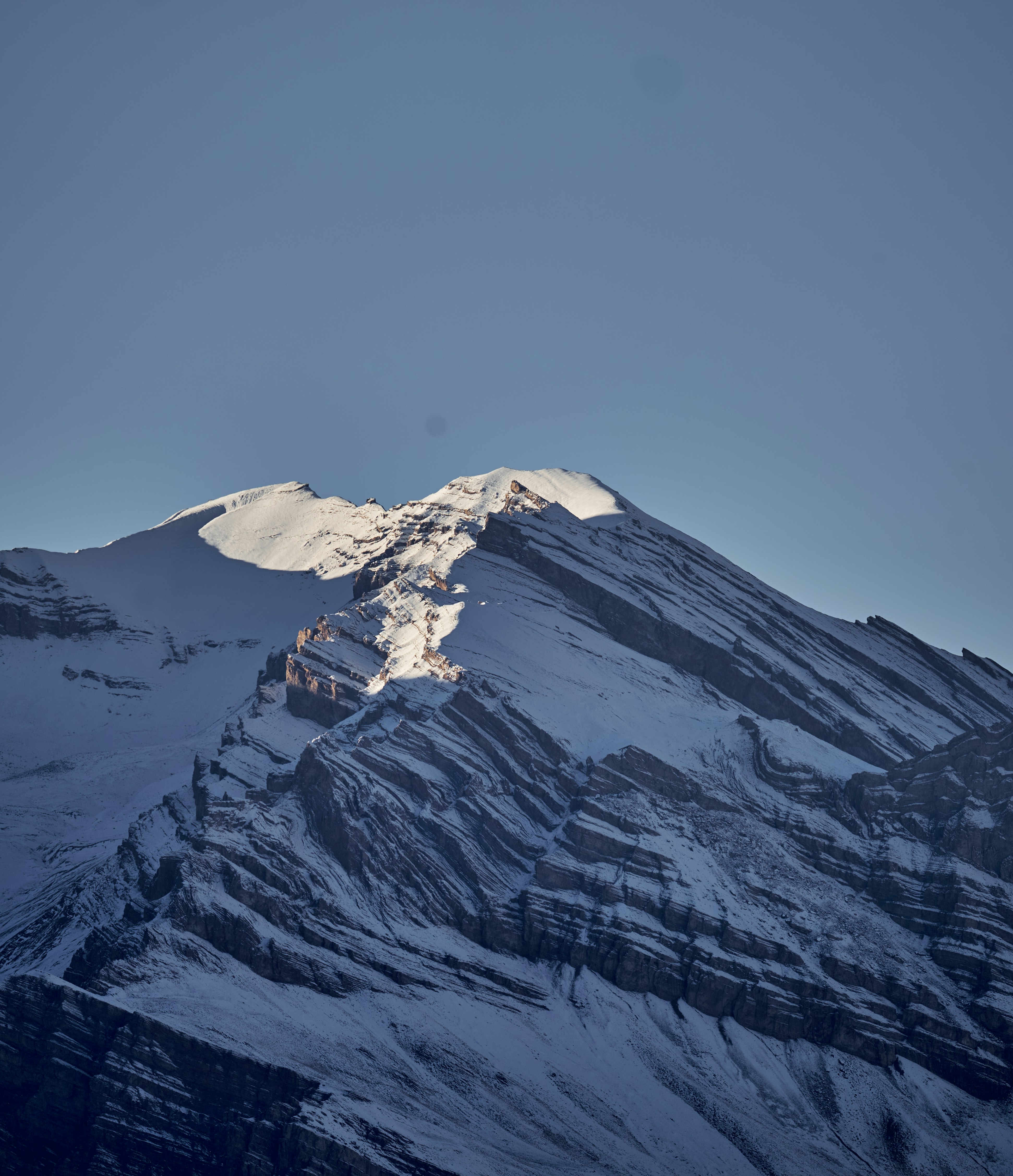 A snow-covered mountain peak with layered rock strata under a clear blue sky
