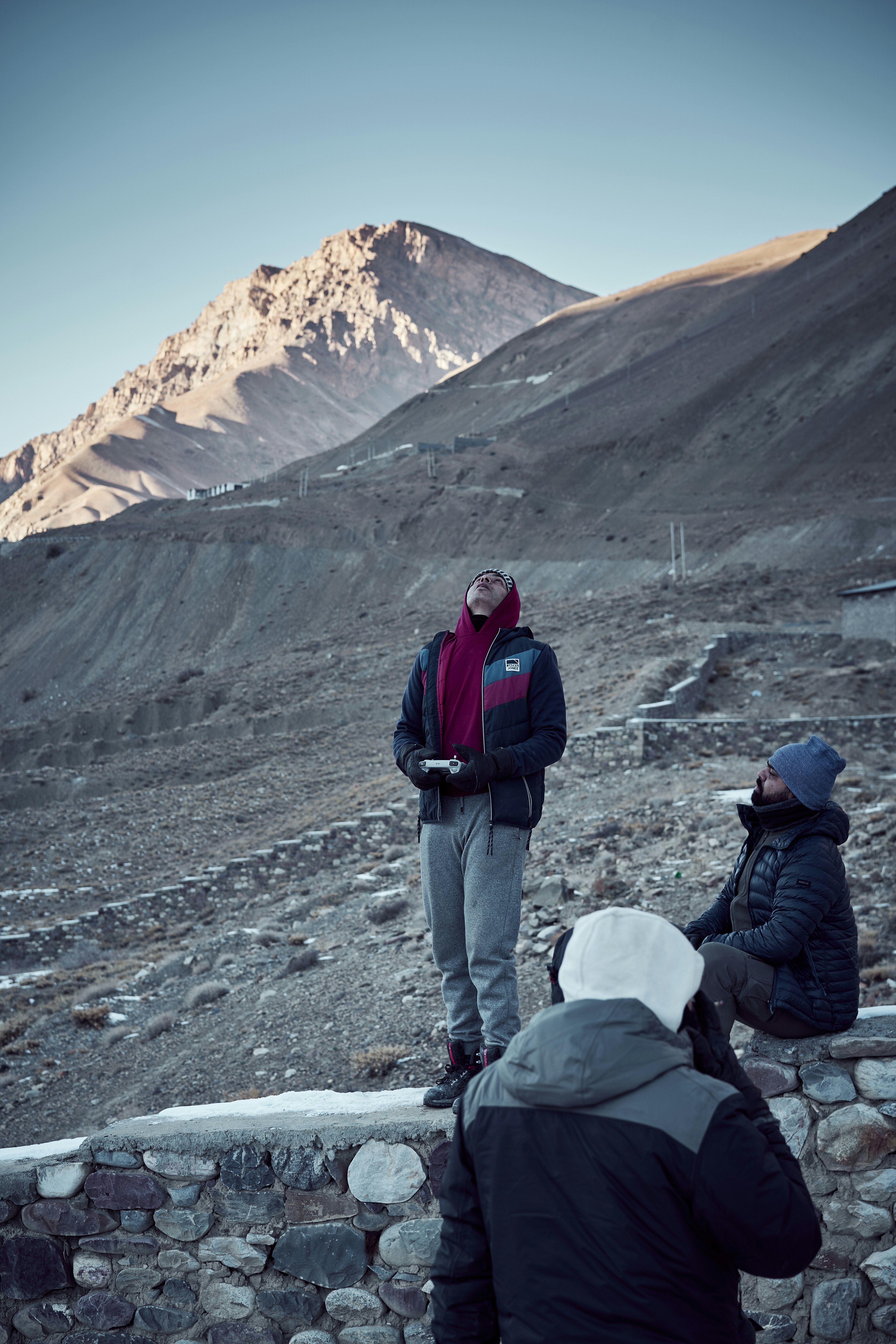 Three people in winter clothing on a rocky mountain slope, one operating a drone