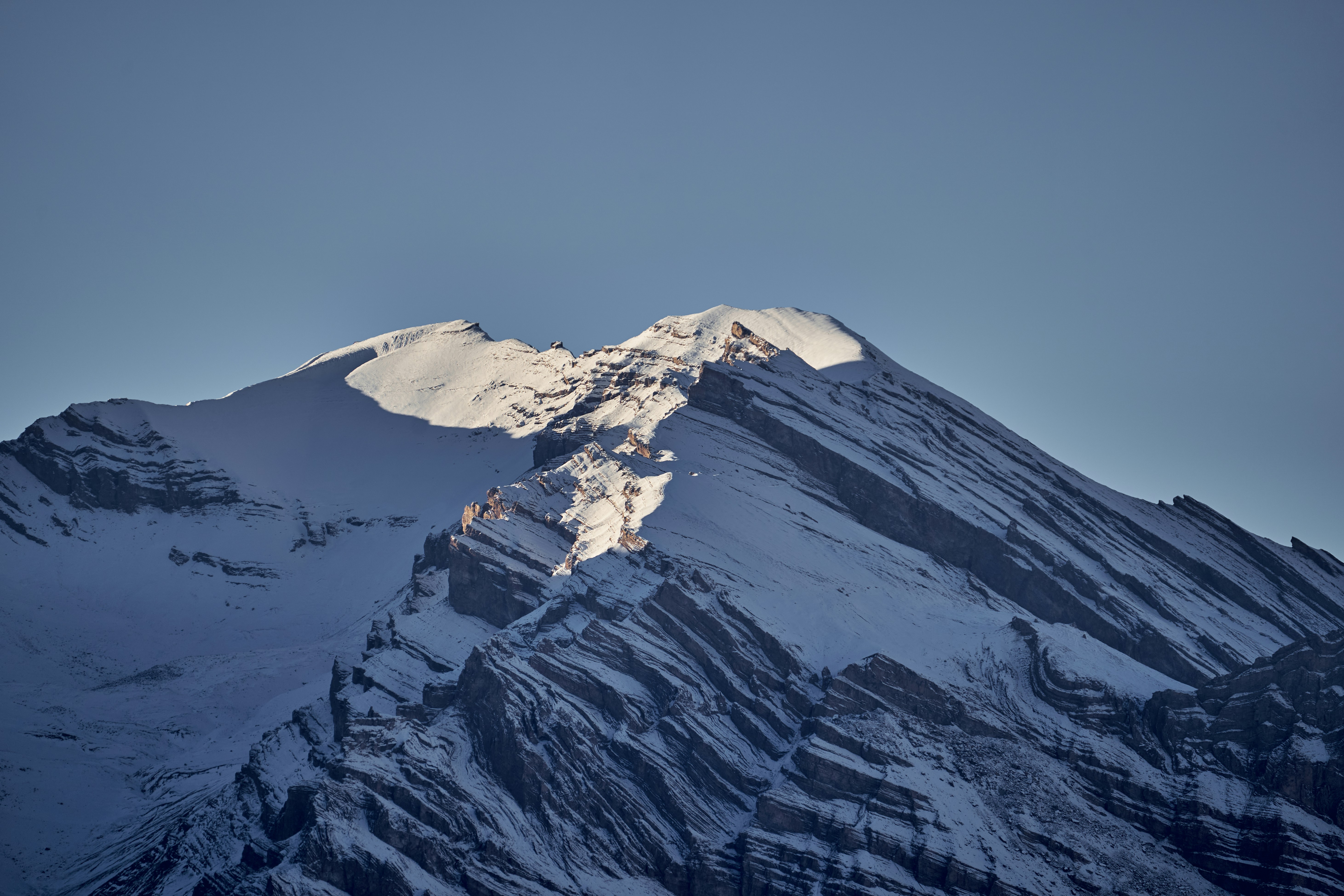 Snow covered mountain peak with jagged rock strata under a clear blue sky