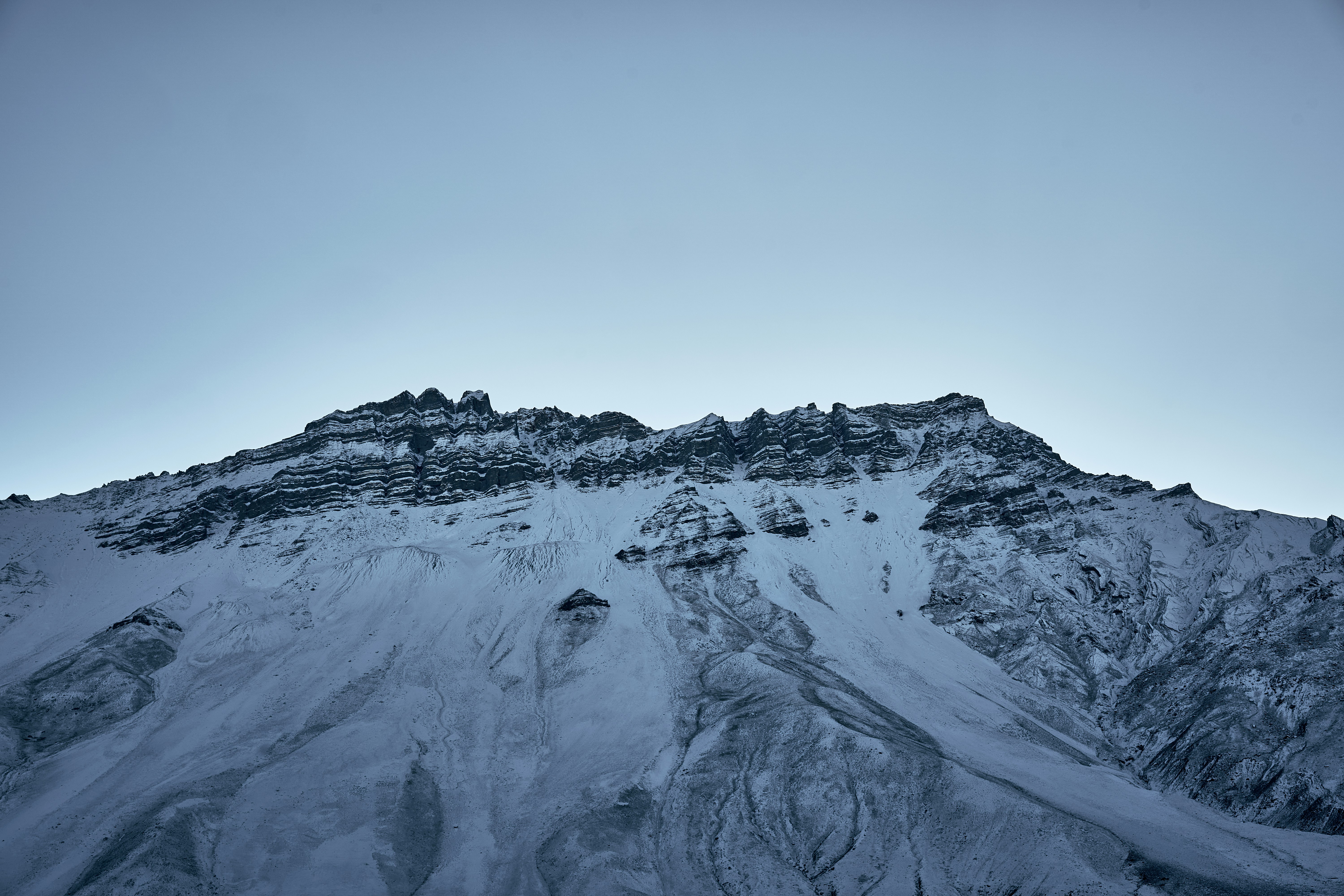 A jagged snow covered mountain peak against a clear pale blue sky