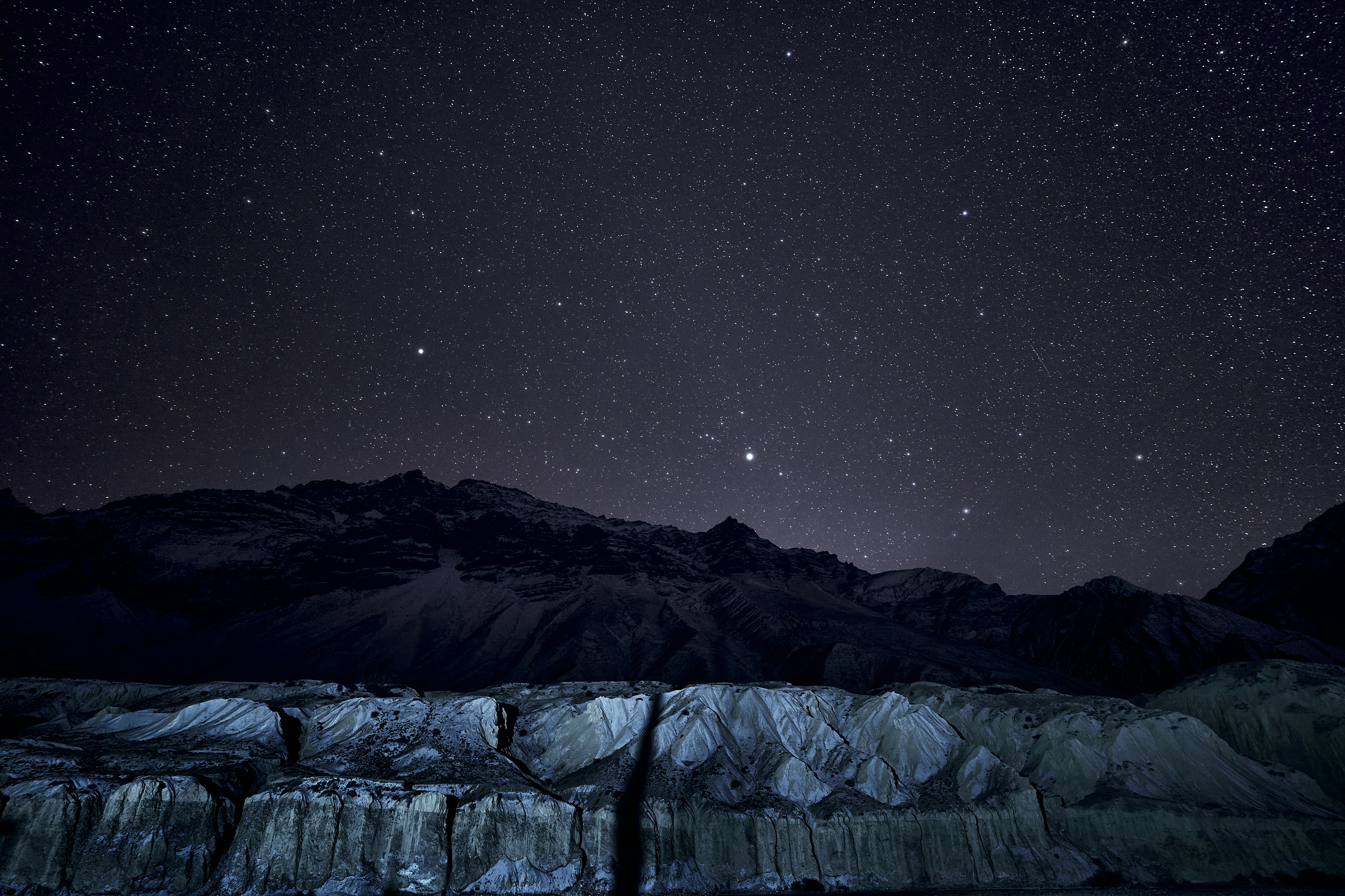 A starry night sky above a dark mountain range and rocky terrain