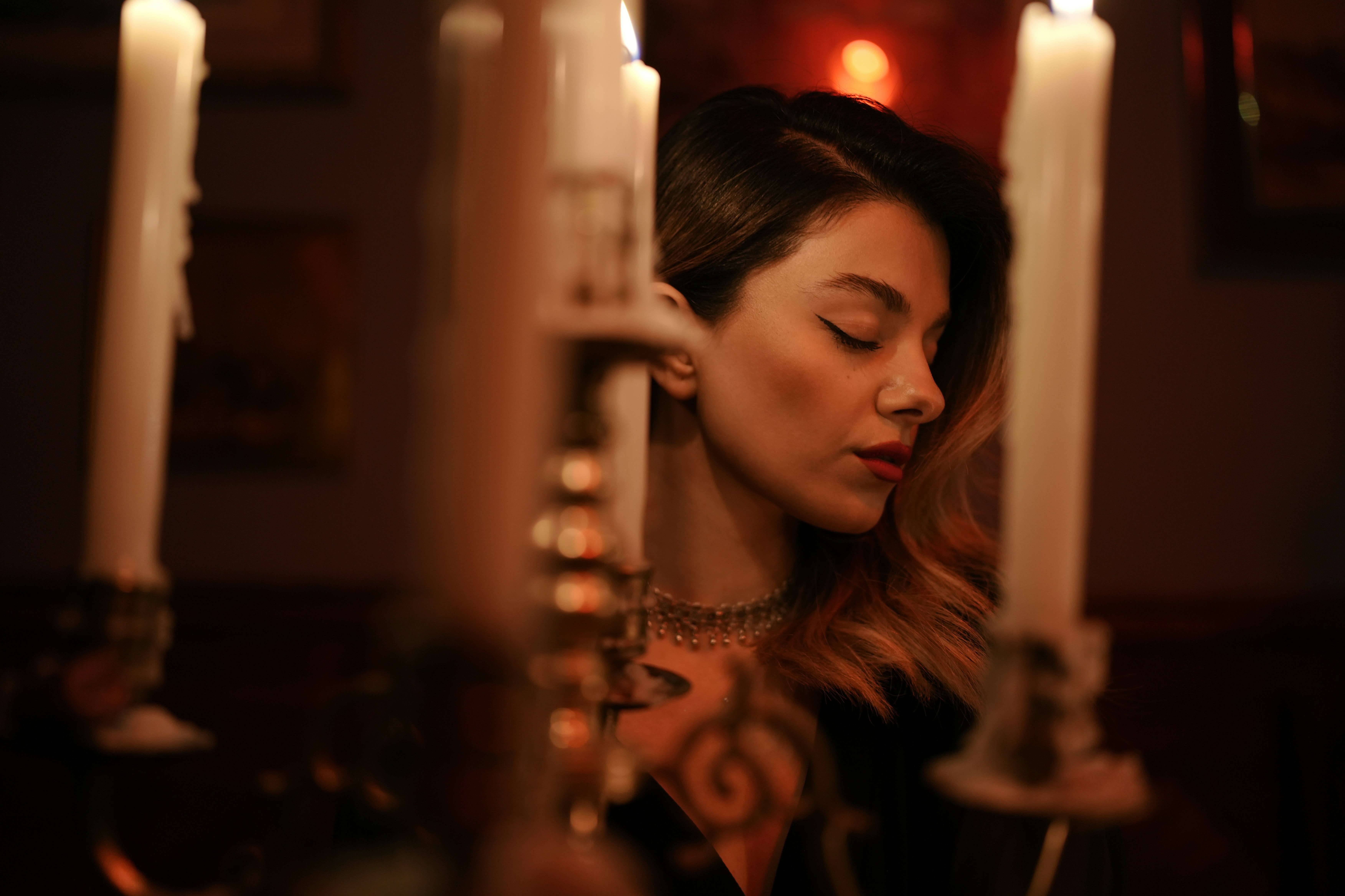 A woman with closed eyes behind tall white candles in a dimly lit room