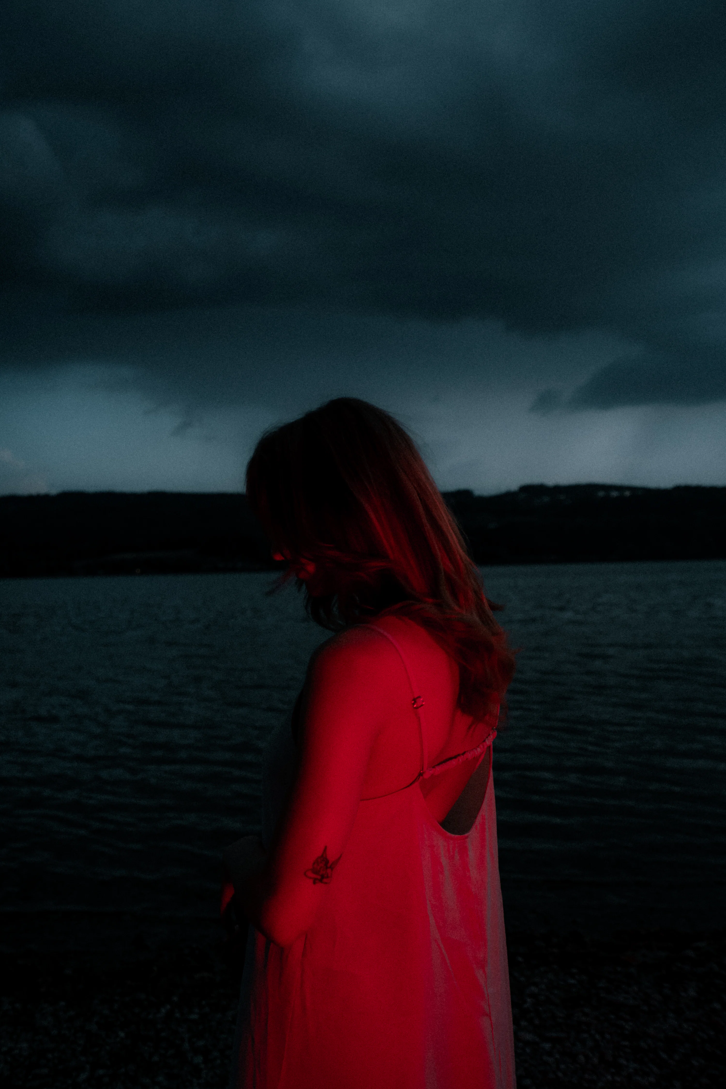 A woman in a light dress illuminated by red light against a dark lake