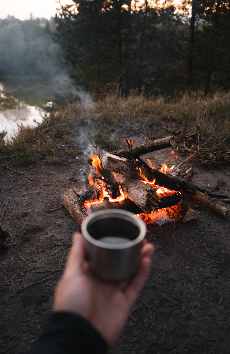 A hand holding a metal mug beside a small campfire on a lakeshore at dusk